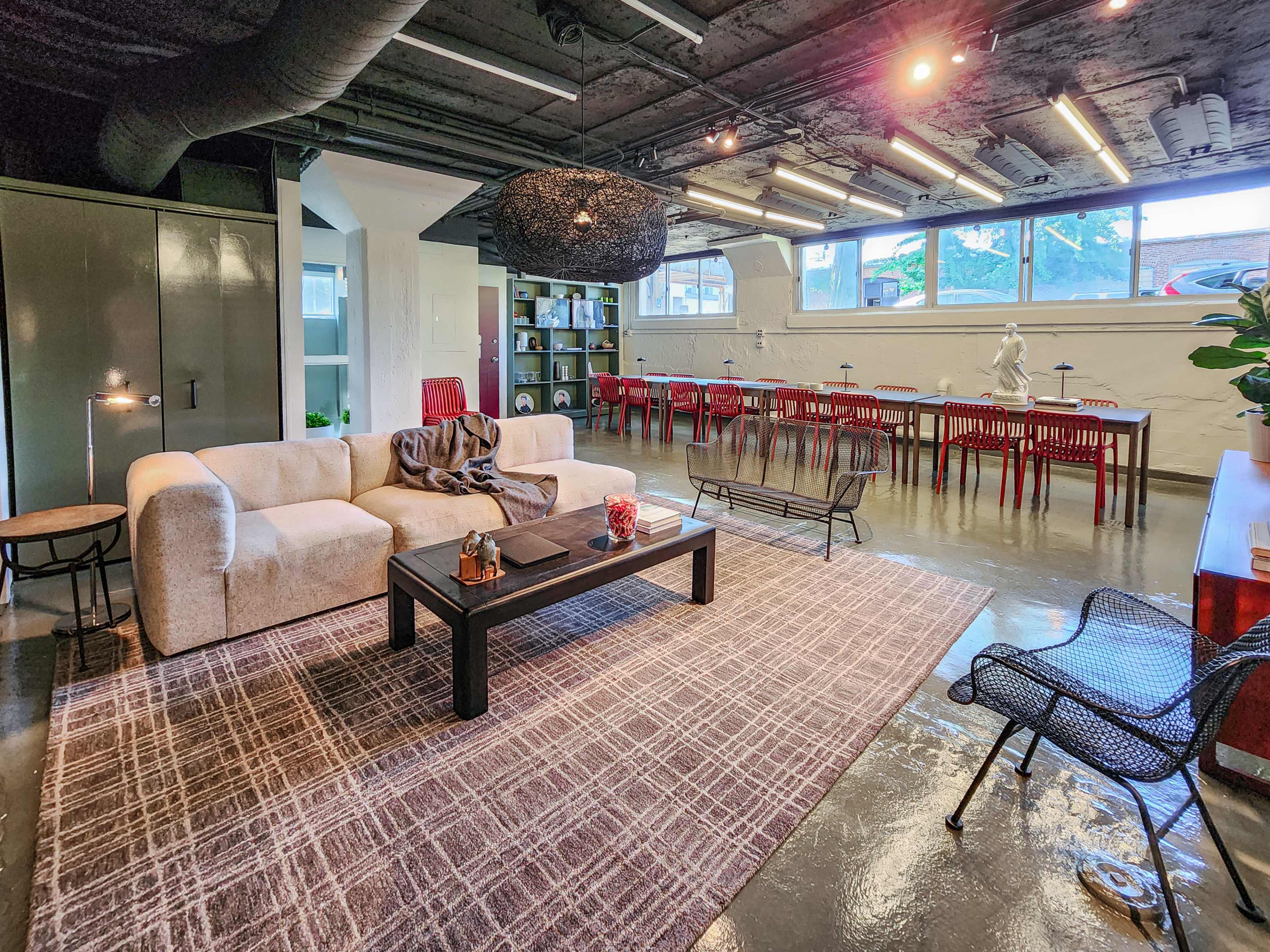 The image shows a modern interior space featuring a cozy living area with a white sofa, a dark coffee table, and a woven light fixture, along with a dining area containing several red chairs and a long table.