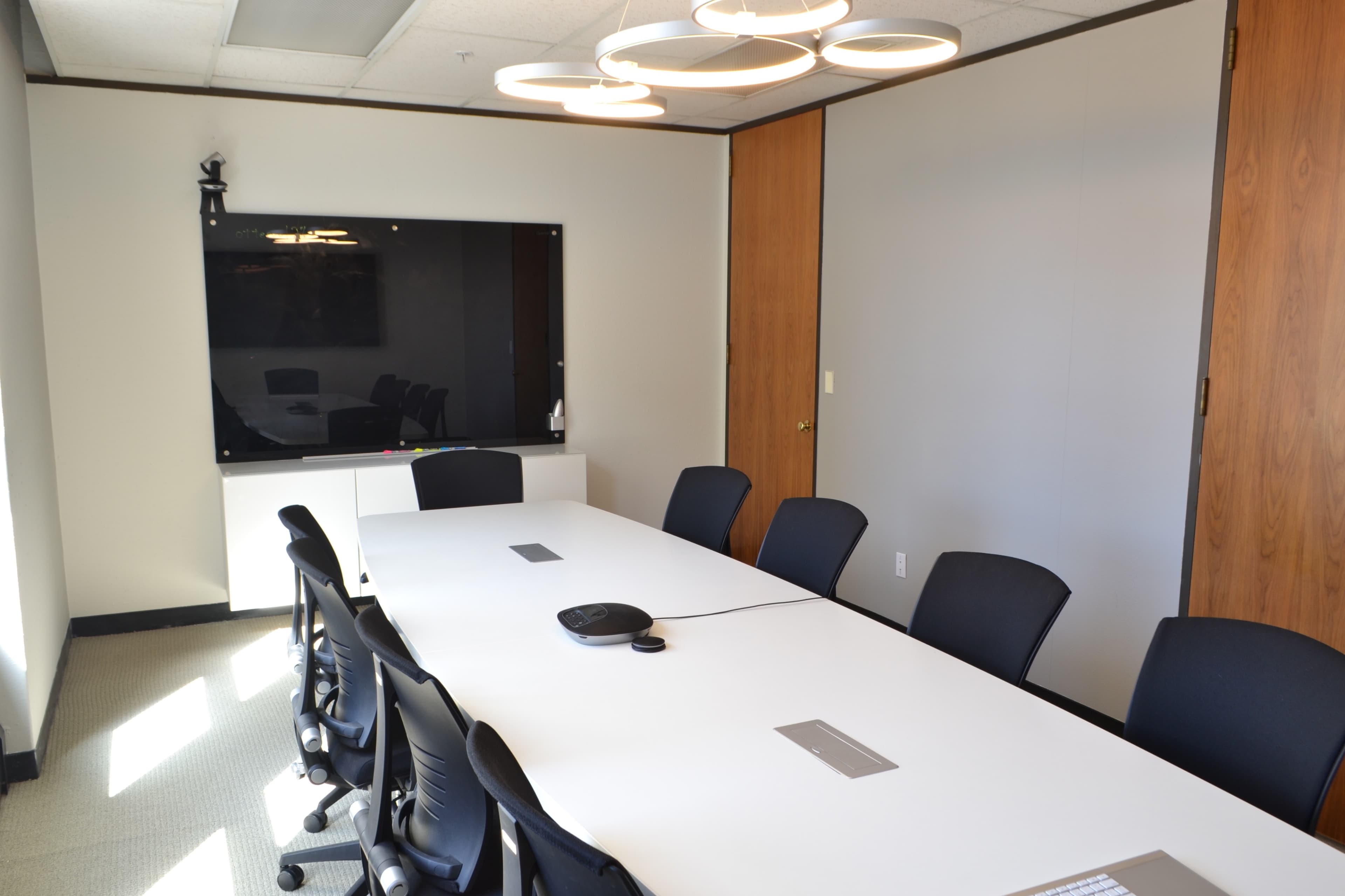 A modern conference room features a large white table surrounded by black chairs, with a flat-screen TV mounted on the wall.