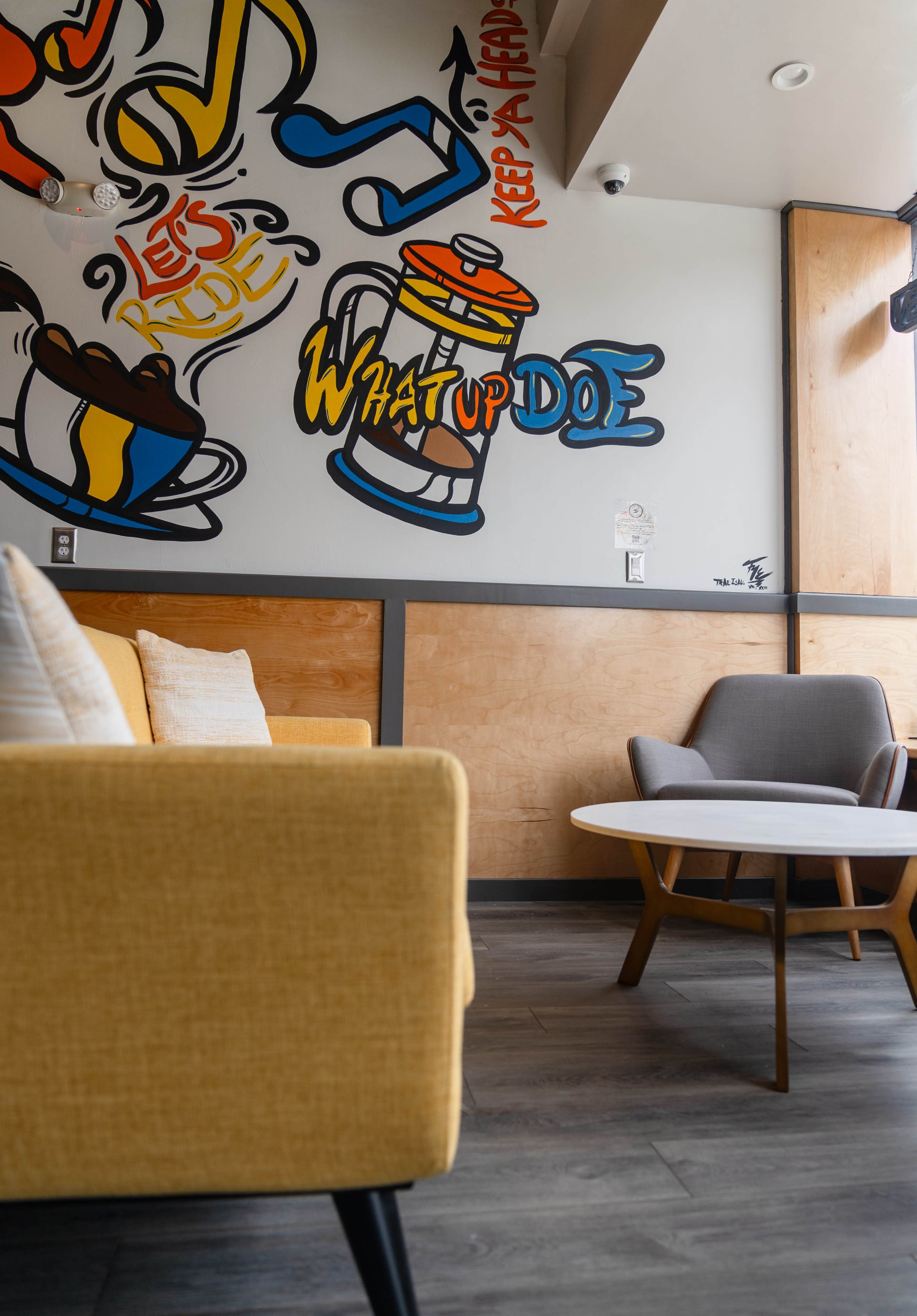 The scene features a cozy seating area with a yellow couch and a gray chair, alongside a round table, set against a colorful mural on the wall with phrases and illustrations.