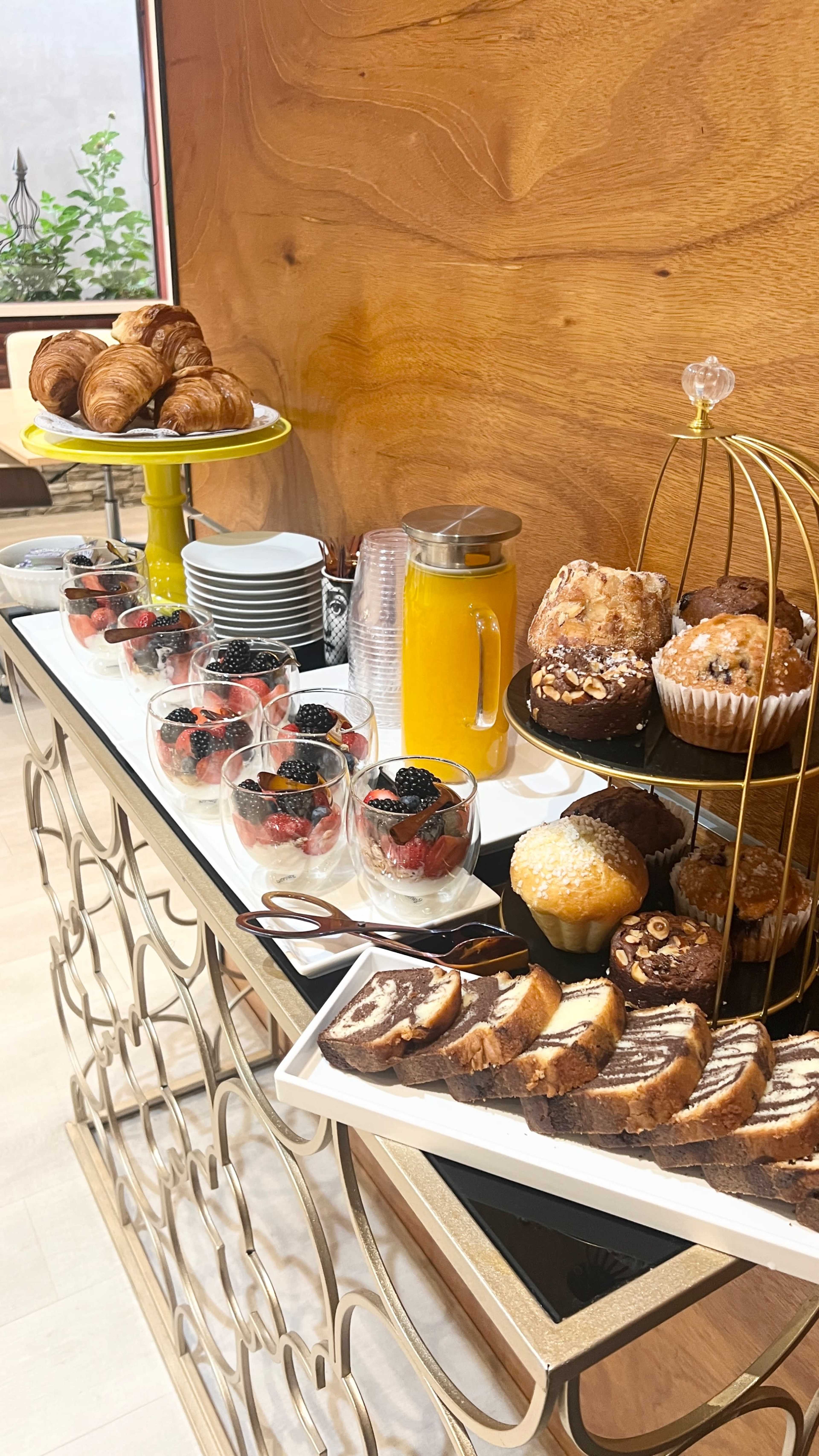 A dessert table features croissants, muffins, chocolate cake, fruit cups with berries, and a pitcher of orange juice.
