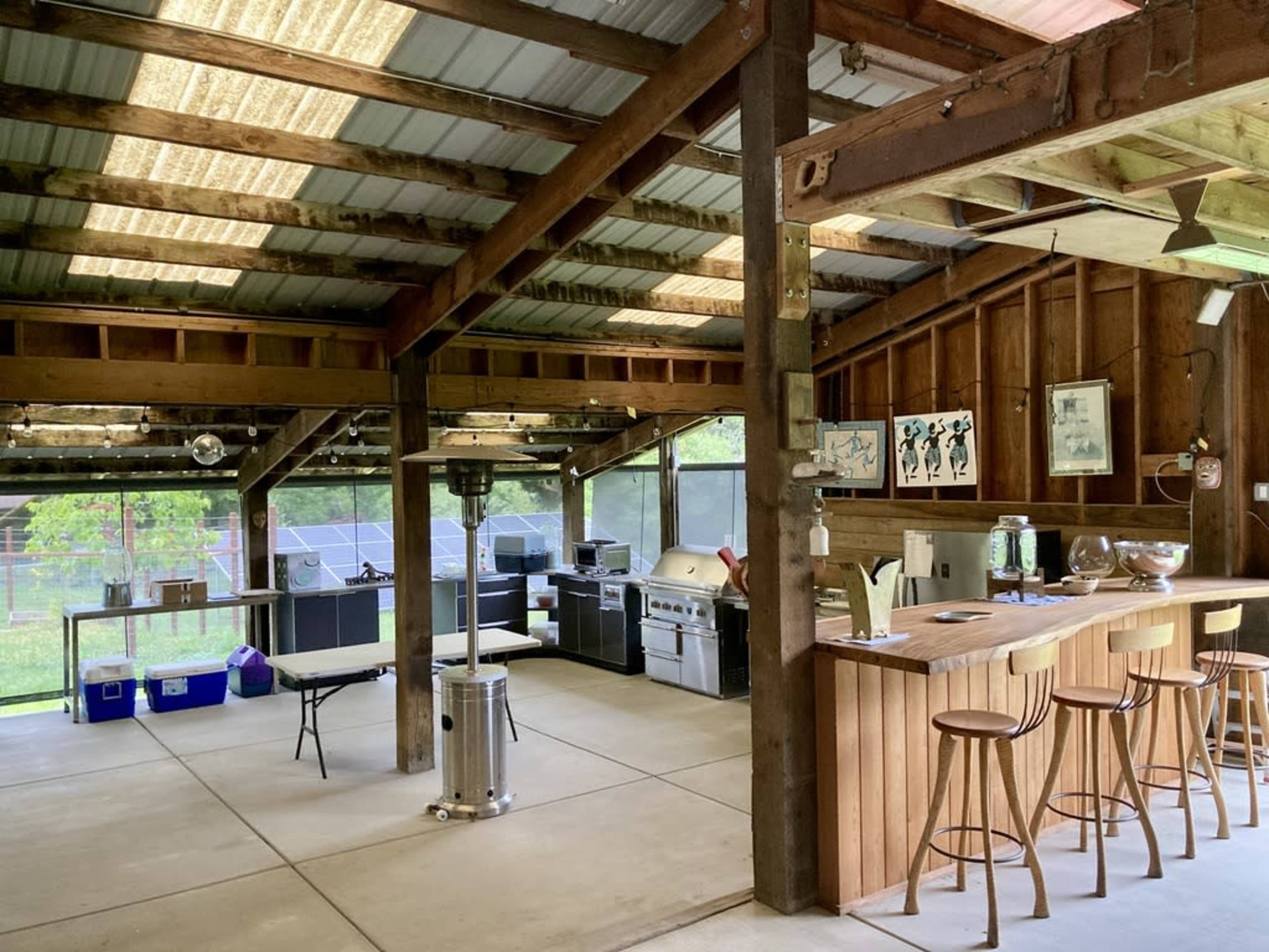 The image shows an open-concept kitchen and dining area in a barn-style structure, featuring wooden beams, bar stools, a grill, and large windows overlooking an outdoor space.