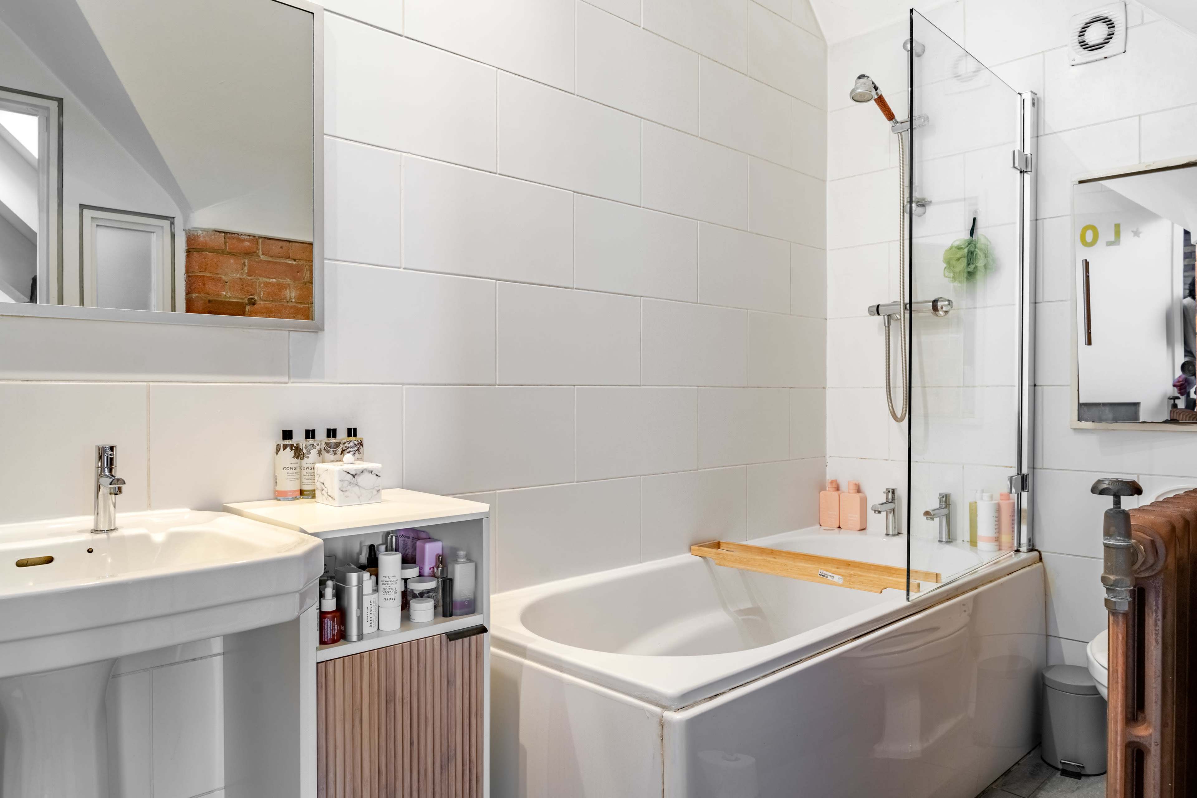 A modern bathroom featuring a white bathtub with a wooden bath rack, a glass shower enclosure, a wall-mounted mirror, and a sink with storage underneath.