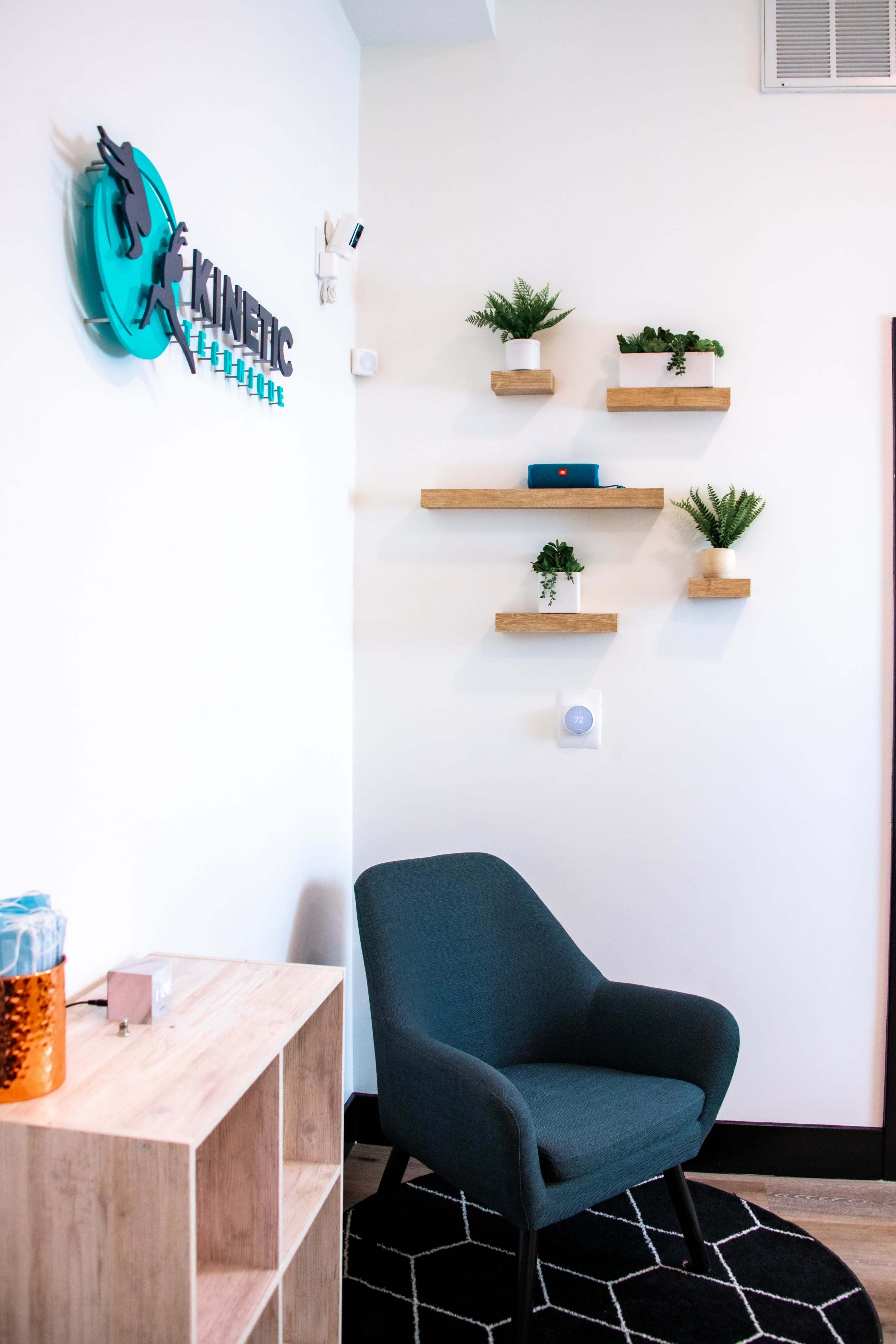 A small reception area featuring a dark green chair, wooden shelves with plants, and a decorative wall sign that reads "KINETIC."