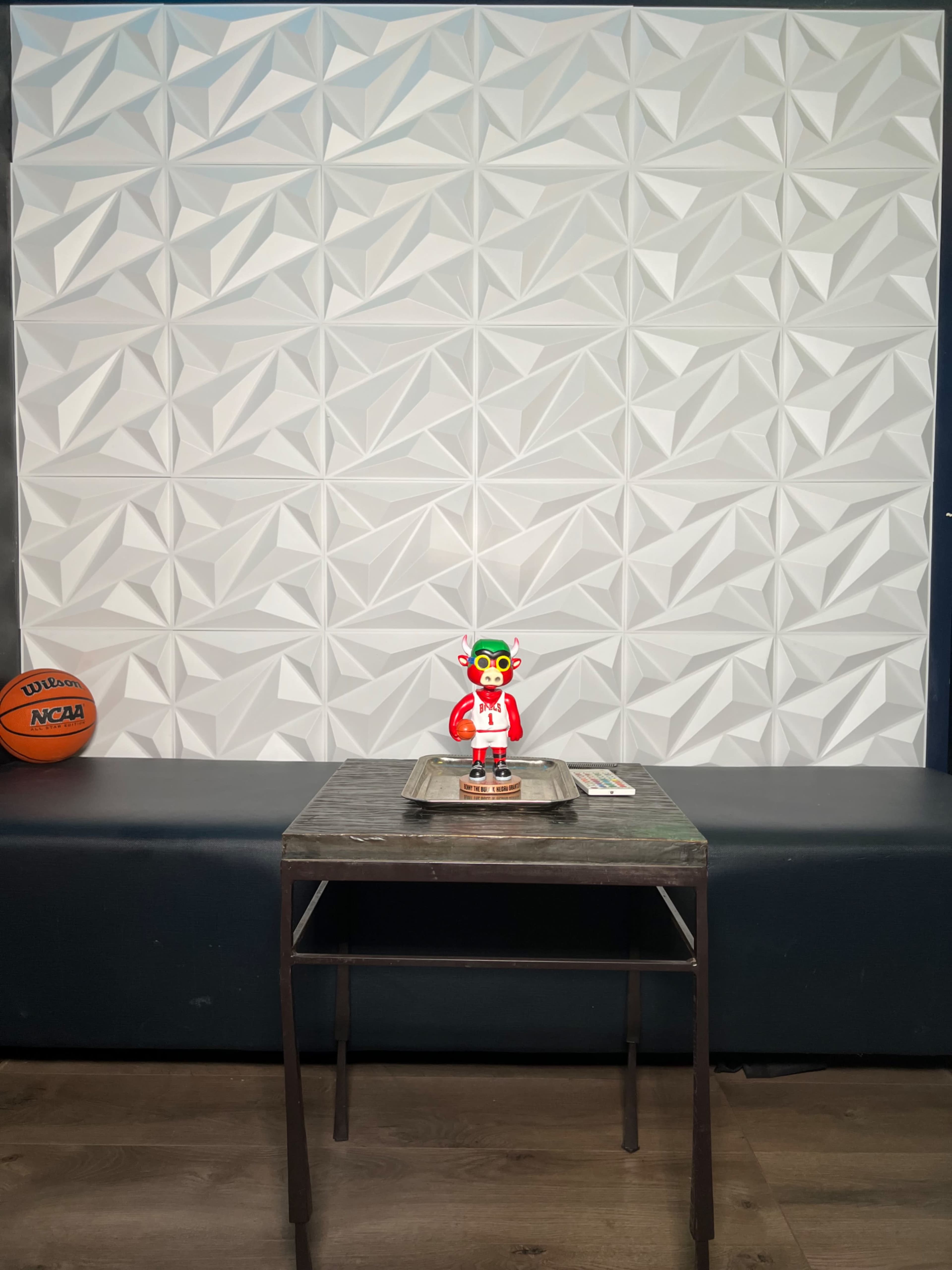 A colorful toy figure stands on a metal table in front of a textured white wall, with a basketball resting on a dark bench to the left.