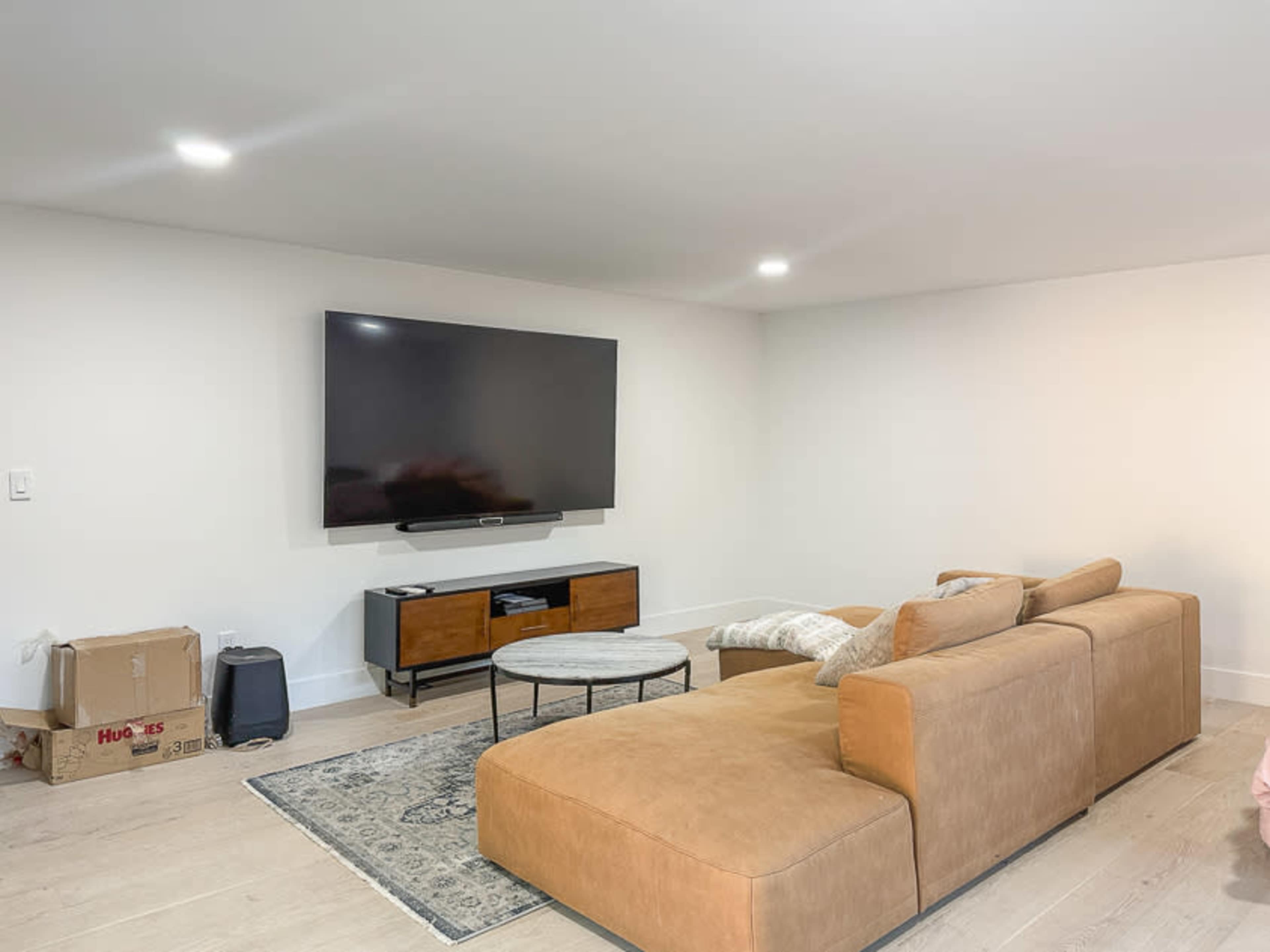 A modern living room features a large flat-screen TV mounted on the wall above a low media console, with a sectional sofa and a coffee table in front.