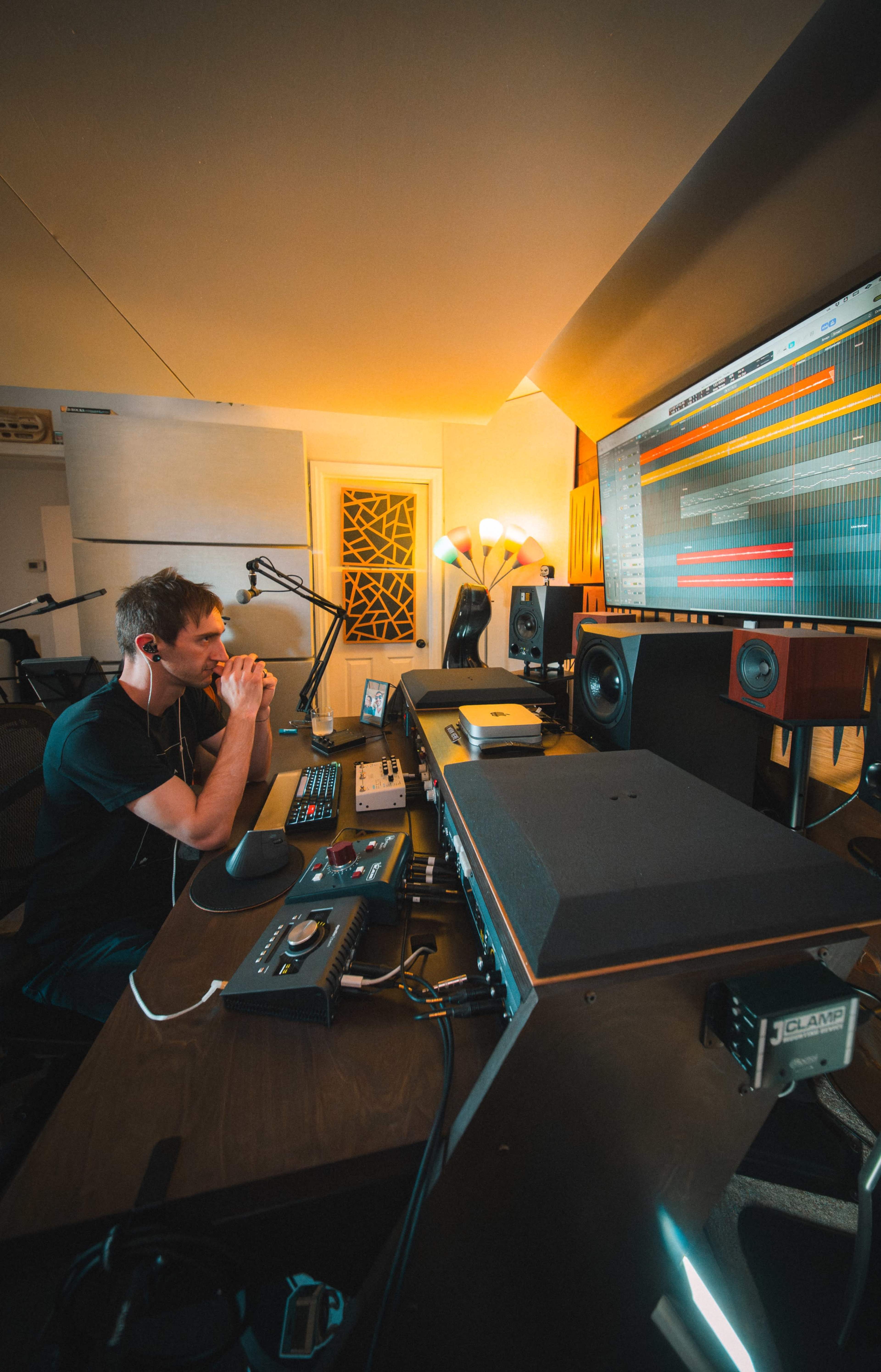 A man sits at a music production workstation, focused on a computer screen displaying audio tracks in a well-lit studio.
