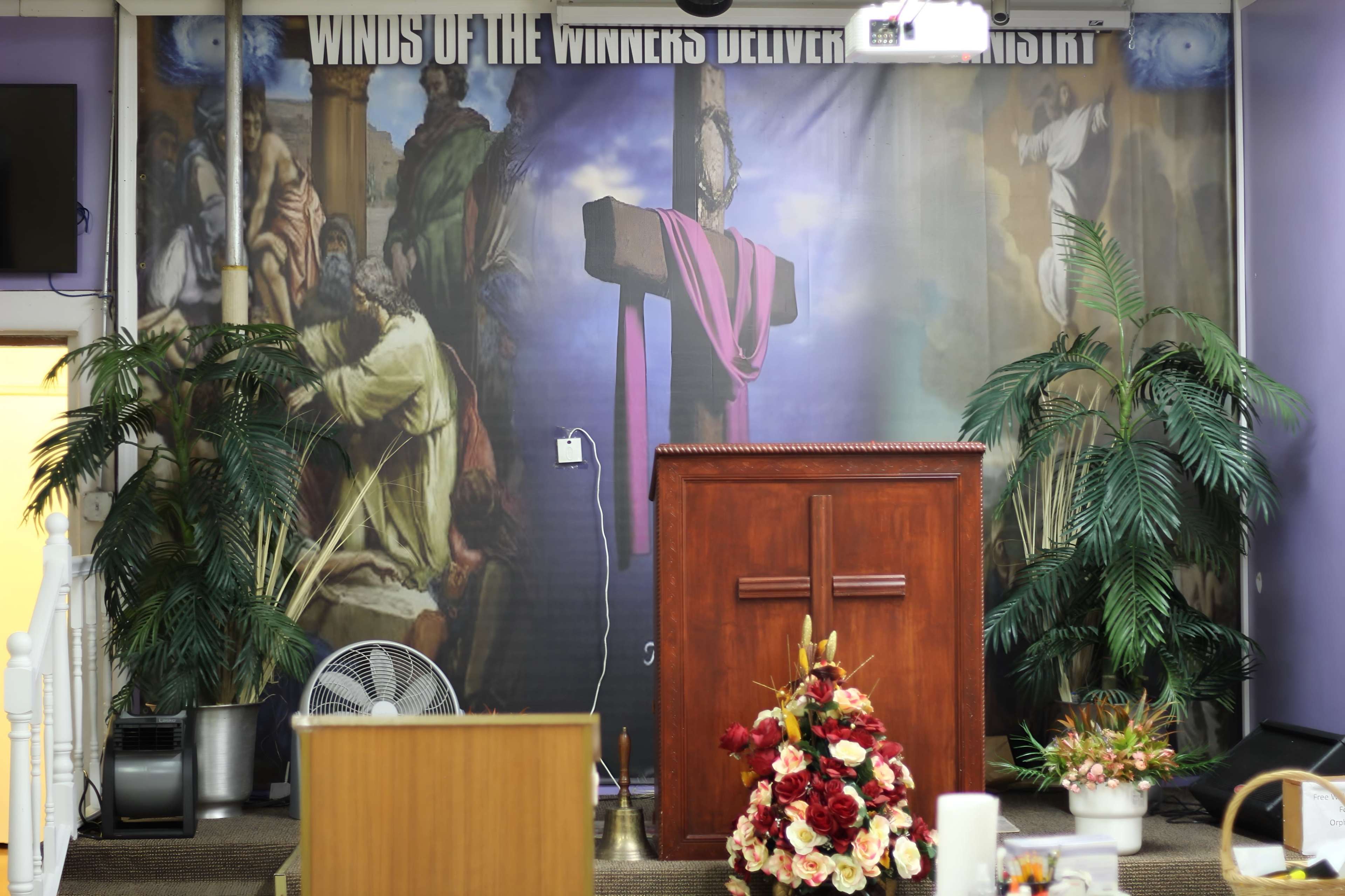 The image shows a church altar with a wooden podium in front of a large mural depicting a crucifixion scene, flanked by potted plants.