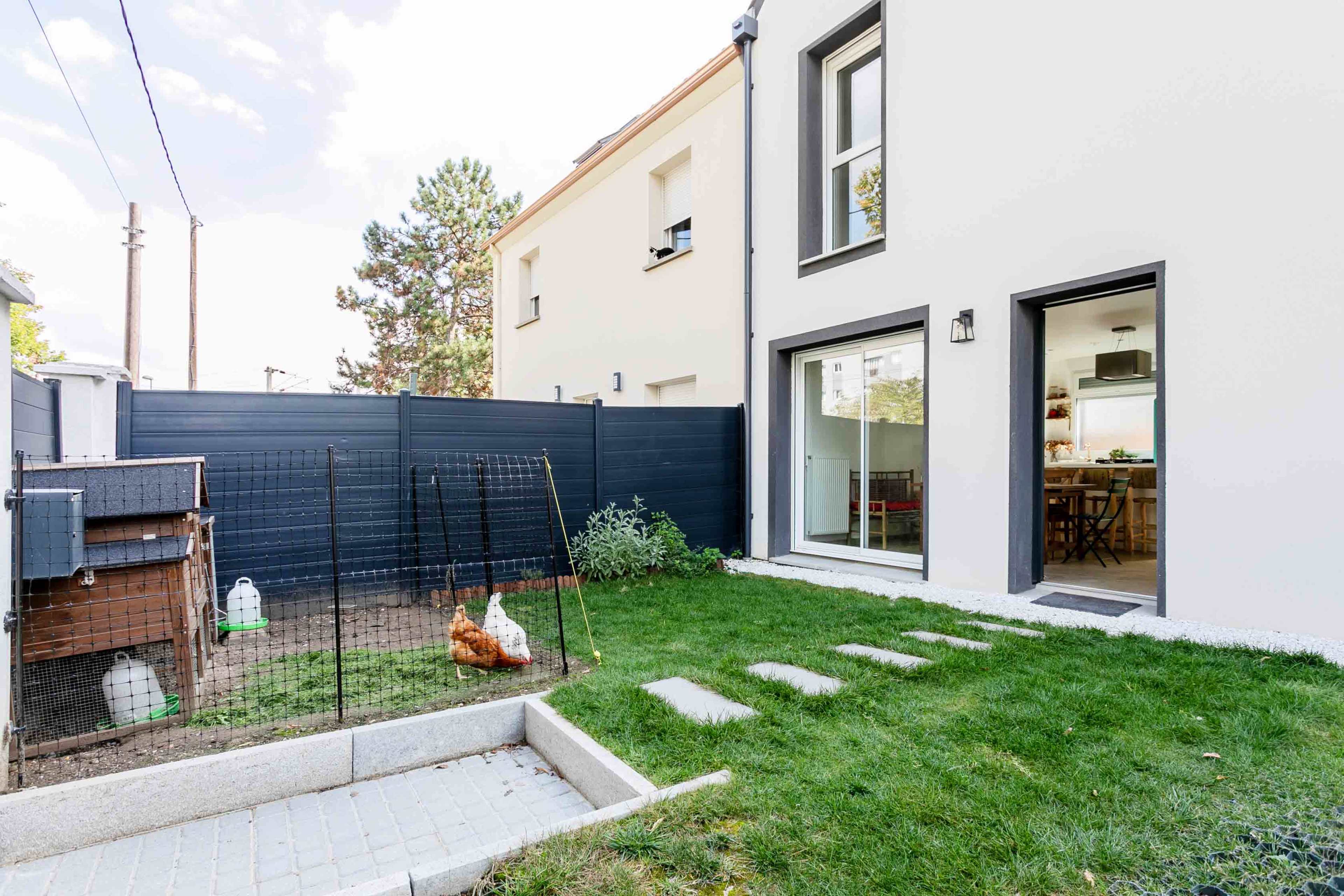 The image shows a backyard with a grassy area, a fence, a small chicken coop, stepping stones leading to a modern house, and a portion of a dining area visible through a glass door.