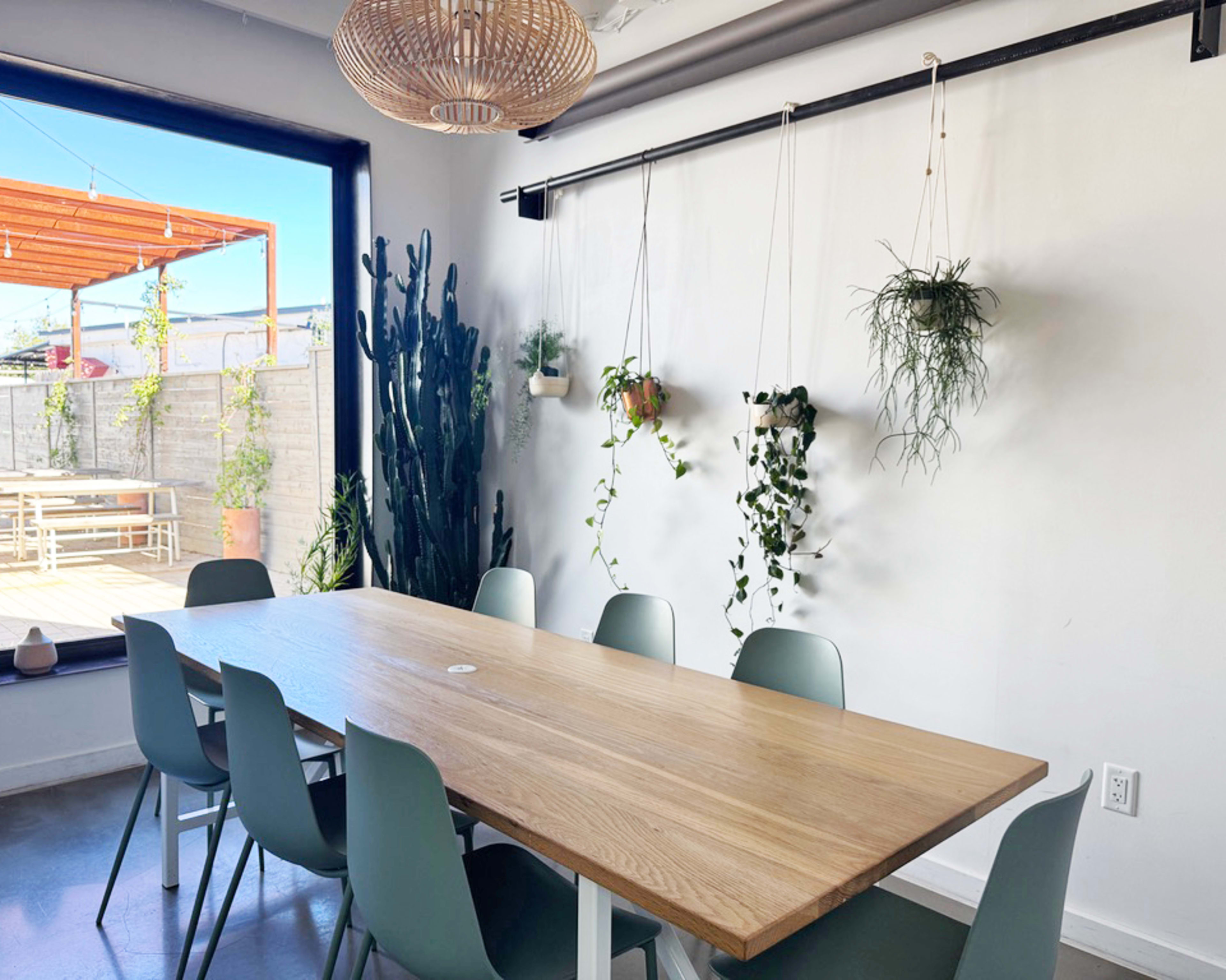 A long wooden table with modern chairs is set in a bright room featuring hanging plants and a large window that opens to an outdoor patio.