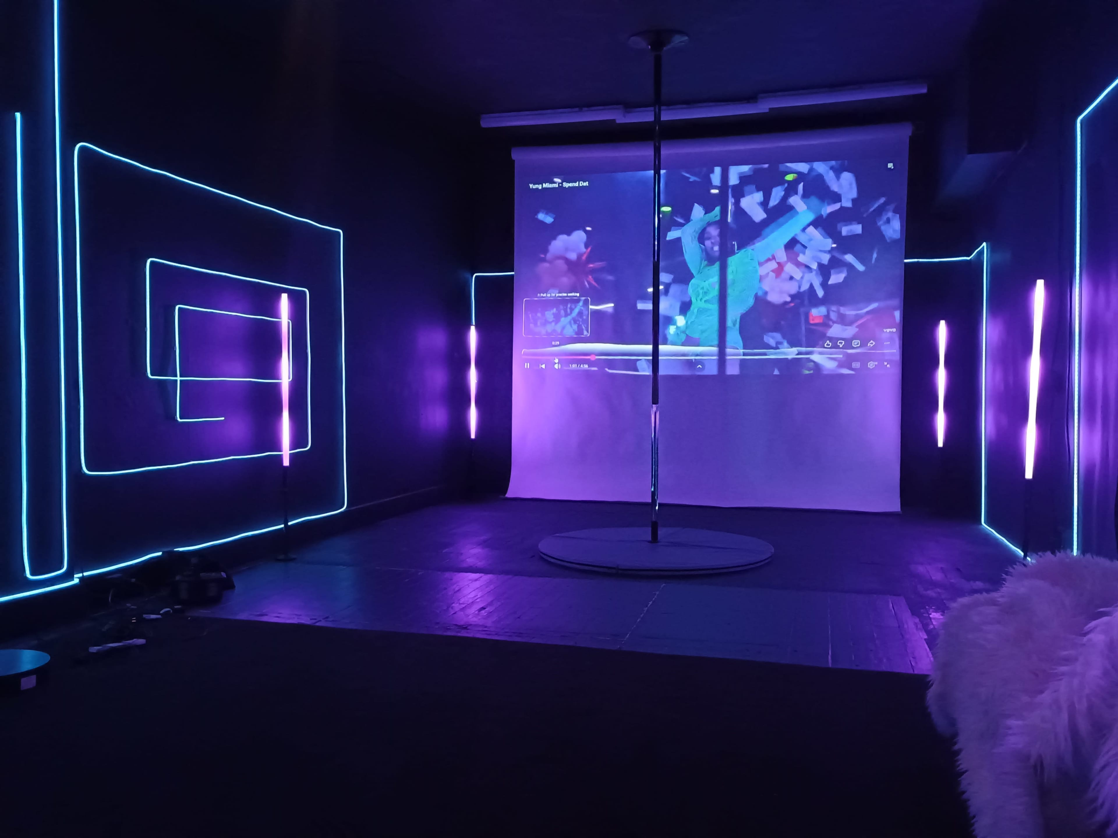 The image shows a dimly lit dance studio featuring a pole in the center, surrounded by neon blue and purple lights along the walls, with a projection screen displaying a video in the background.