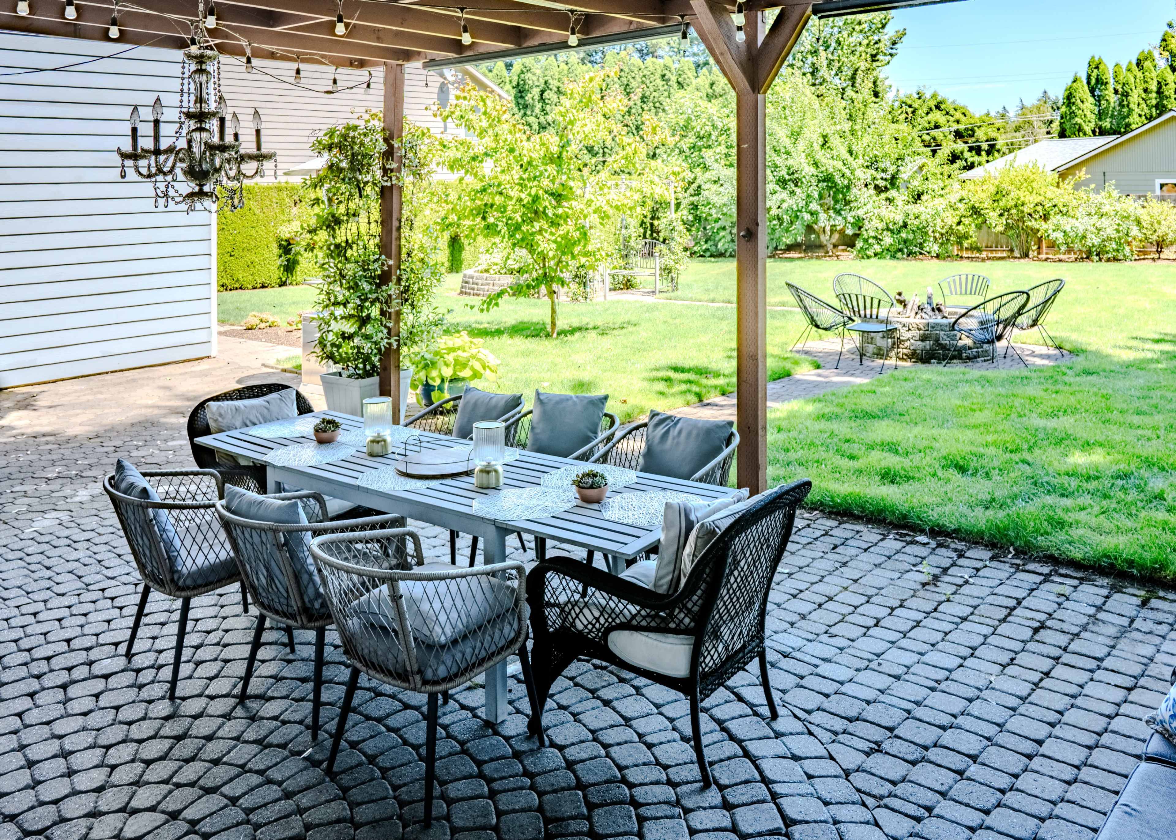 An outdoor dining area is set up under a covered patio, featuring a long table surrounded by chairs and a view of a grassy backyard with additional seating in the distance.