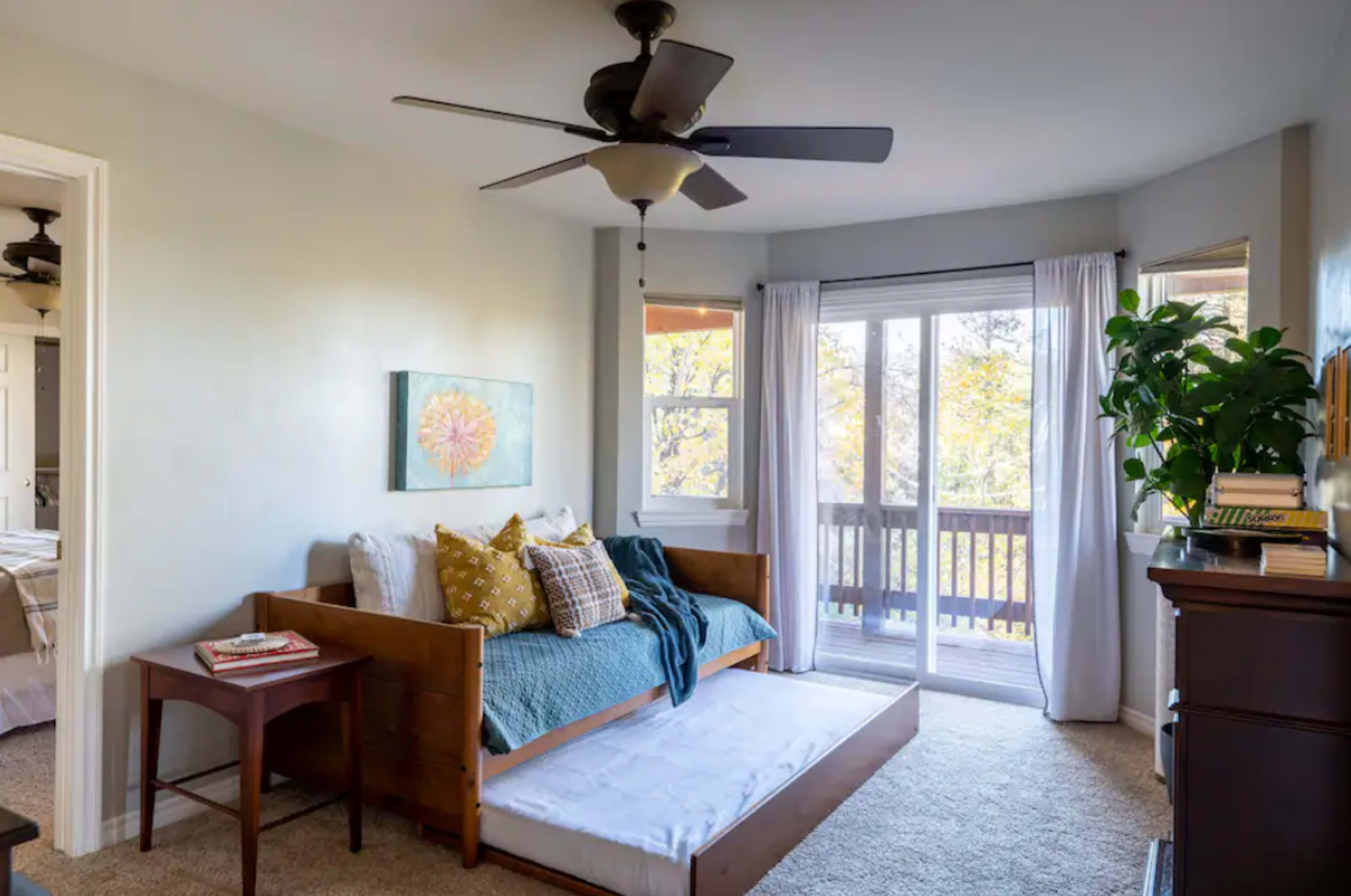 A cozy bedroom features a daybed with decorative pillows, a wooden side table, and large windows overlooking a balcony with greenery.