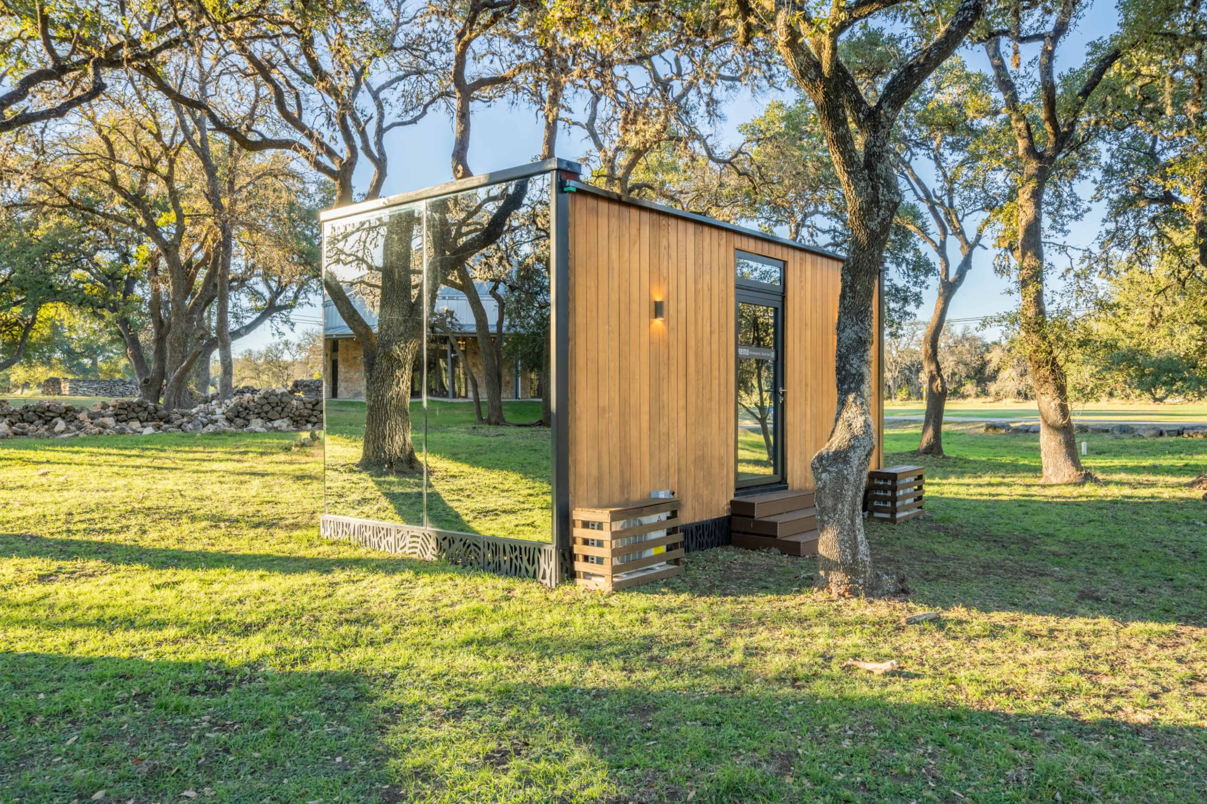 A modern, rectangular cabin with wooden siding and mirrored glass is situated among trees on a grassy landscape.