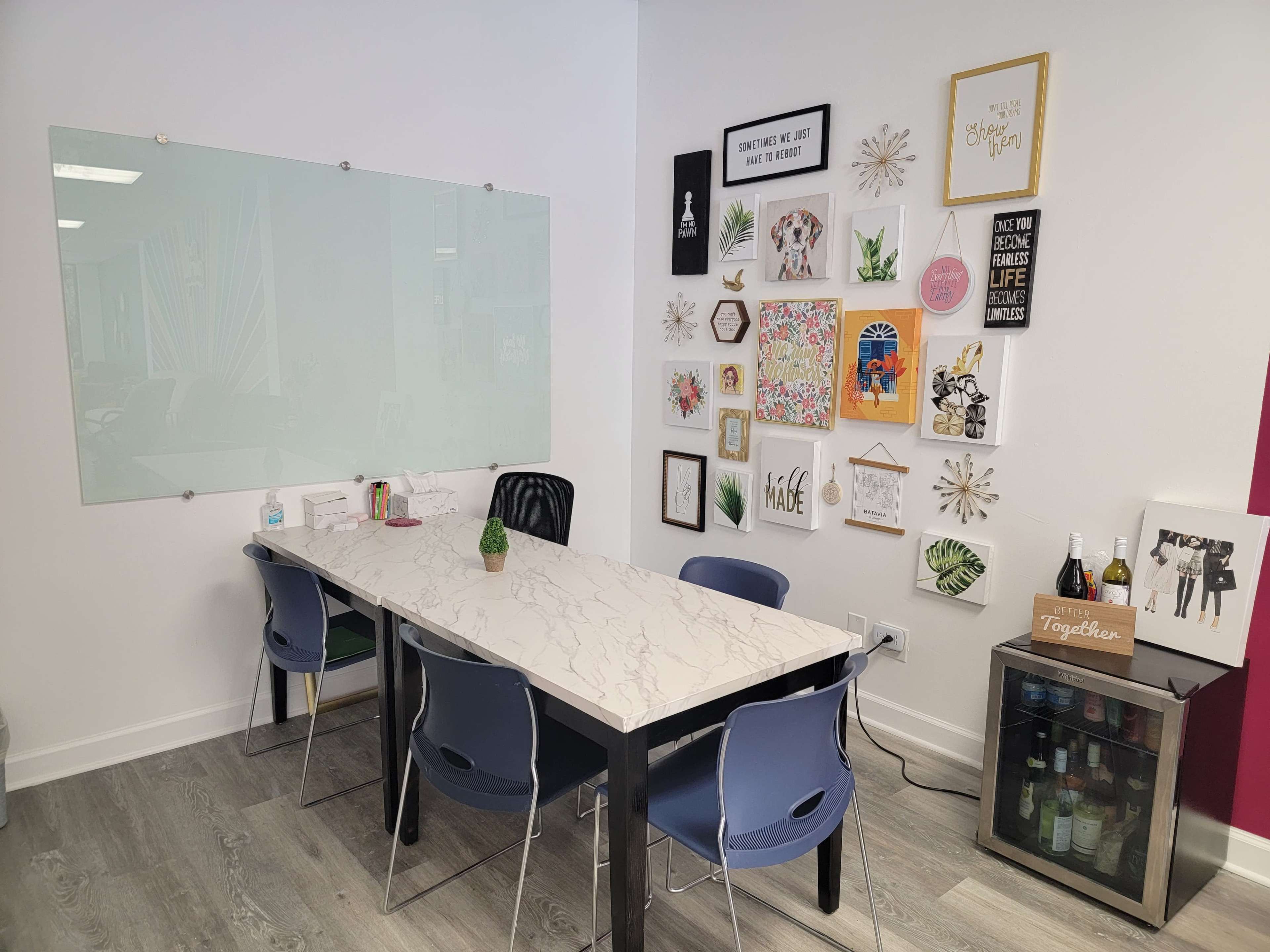 A modern meeting room with a marble table, blue chairs, a glass whiteboard, and a wall decorated with various framed artworks and a small beverage refrigerator.