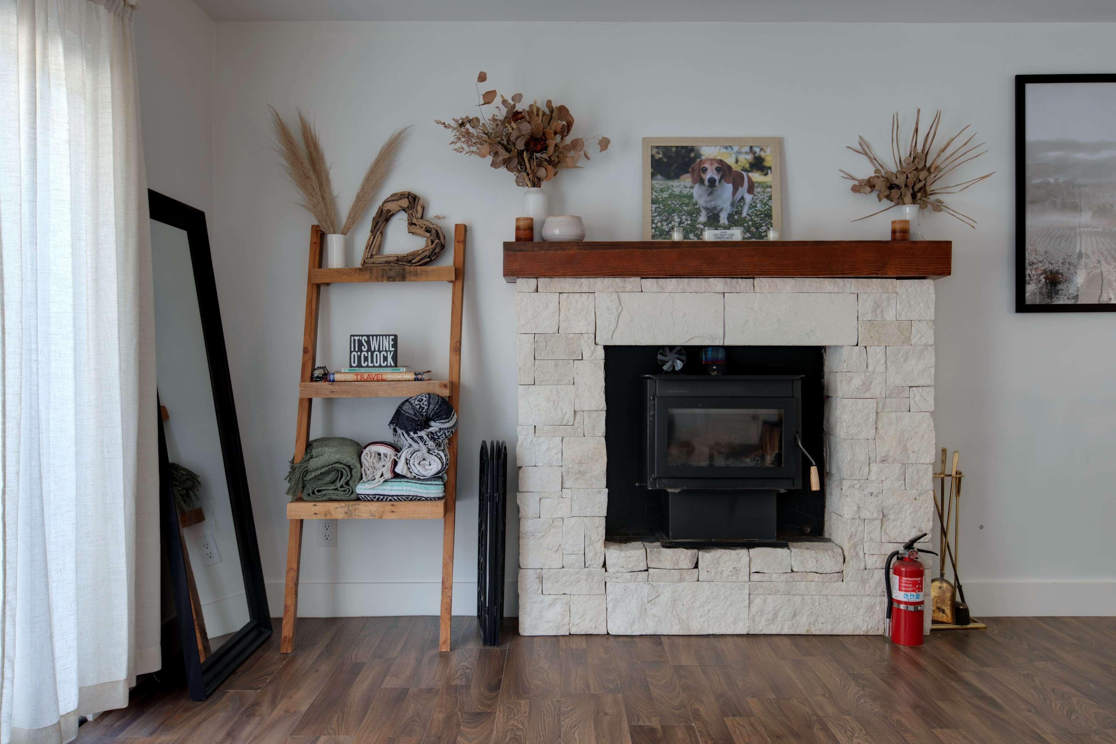 The room features a stone fireplace with a wooden mantel, flanked by a leaning ladder shelf displaying blankets and a large mirror.