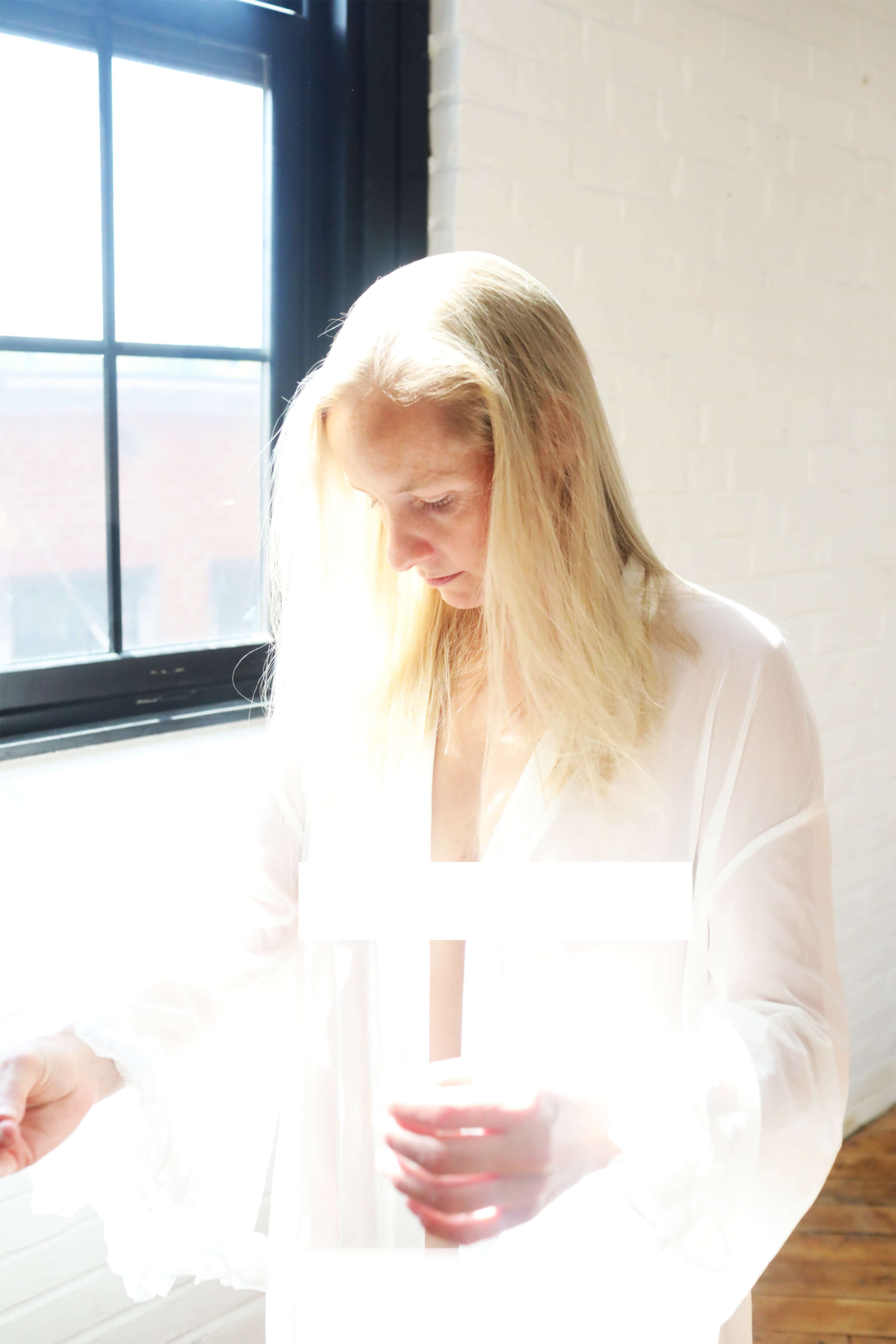 A woman with long blond hair stands in a sunlit room, wearing a white sheer blouse and looking down at her hands.