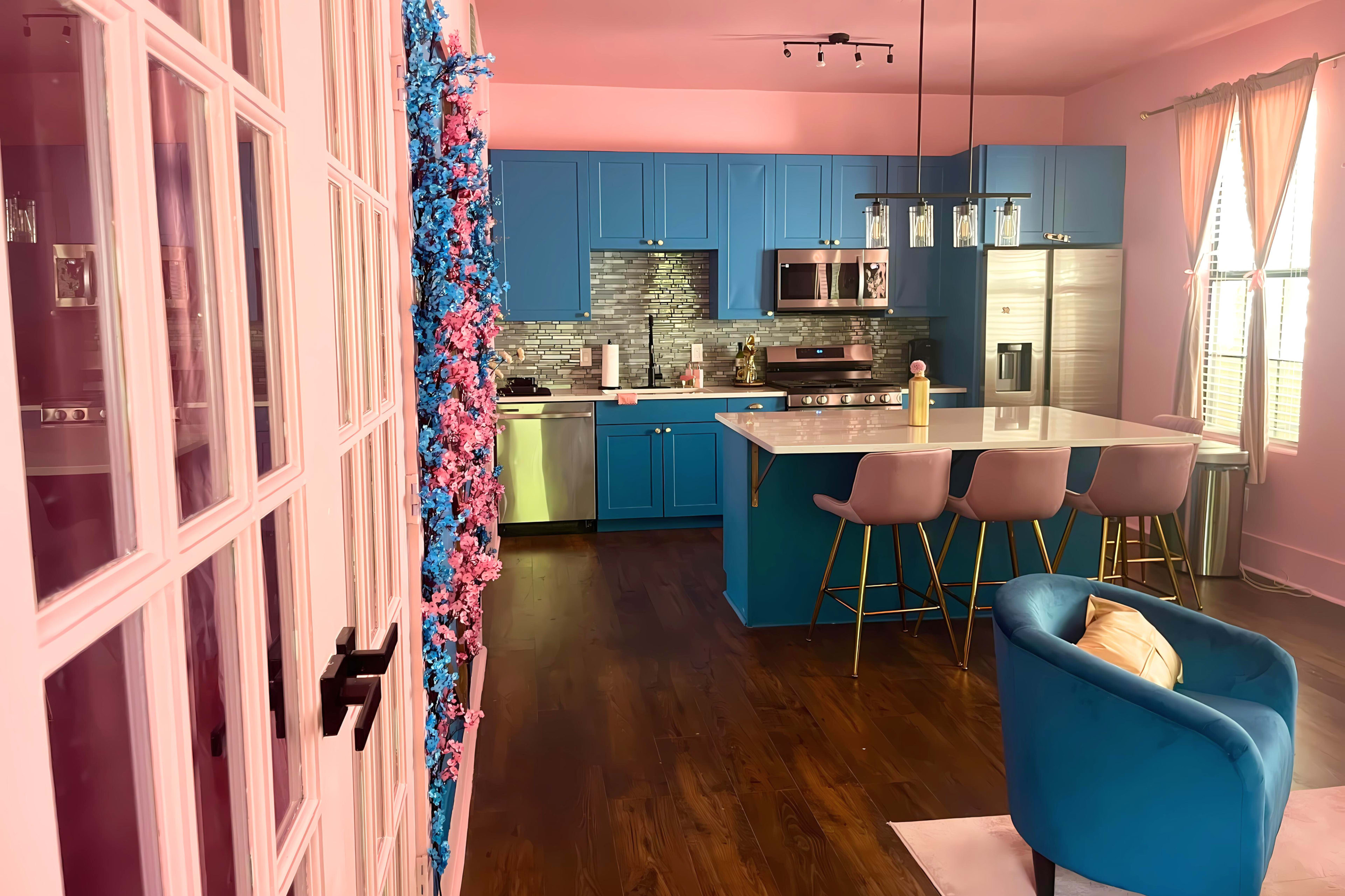 A kitchen with blue cabinetry, a white island, and beige bar stools, adjacent to a living area with a teal chair and a pink wall.