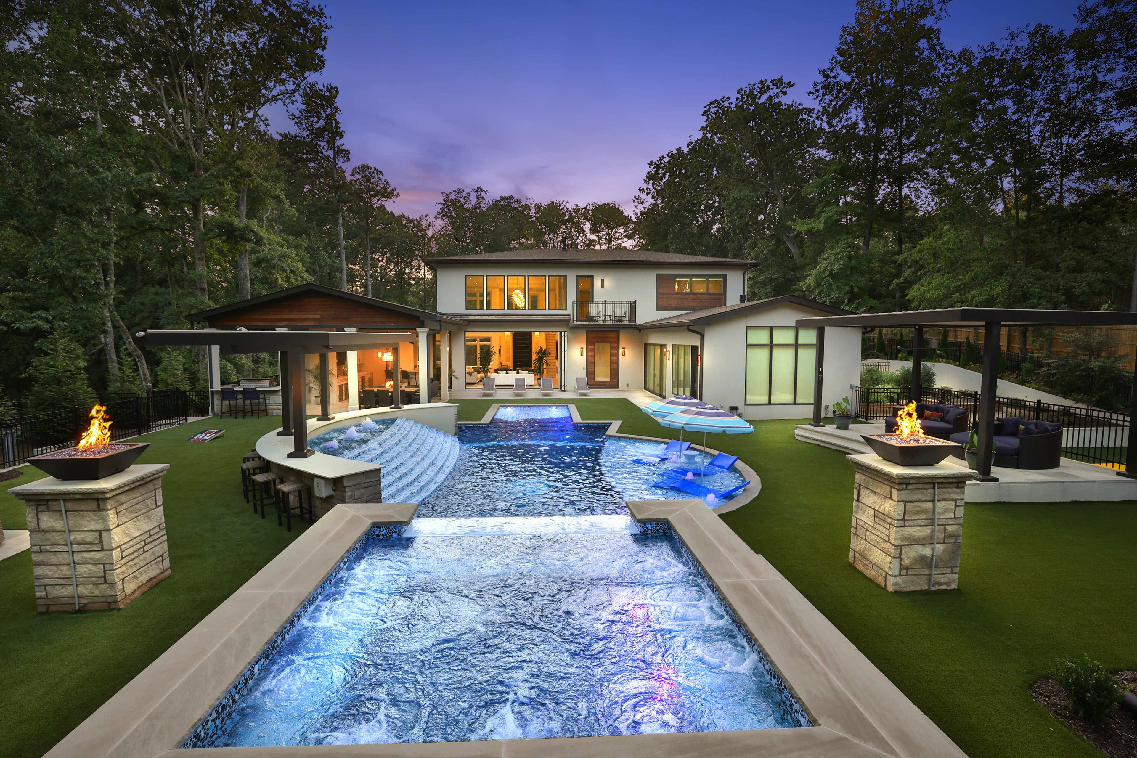 A modern house with a large swimming pool and outdoor seating area, surrounded by trees at dusk.