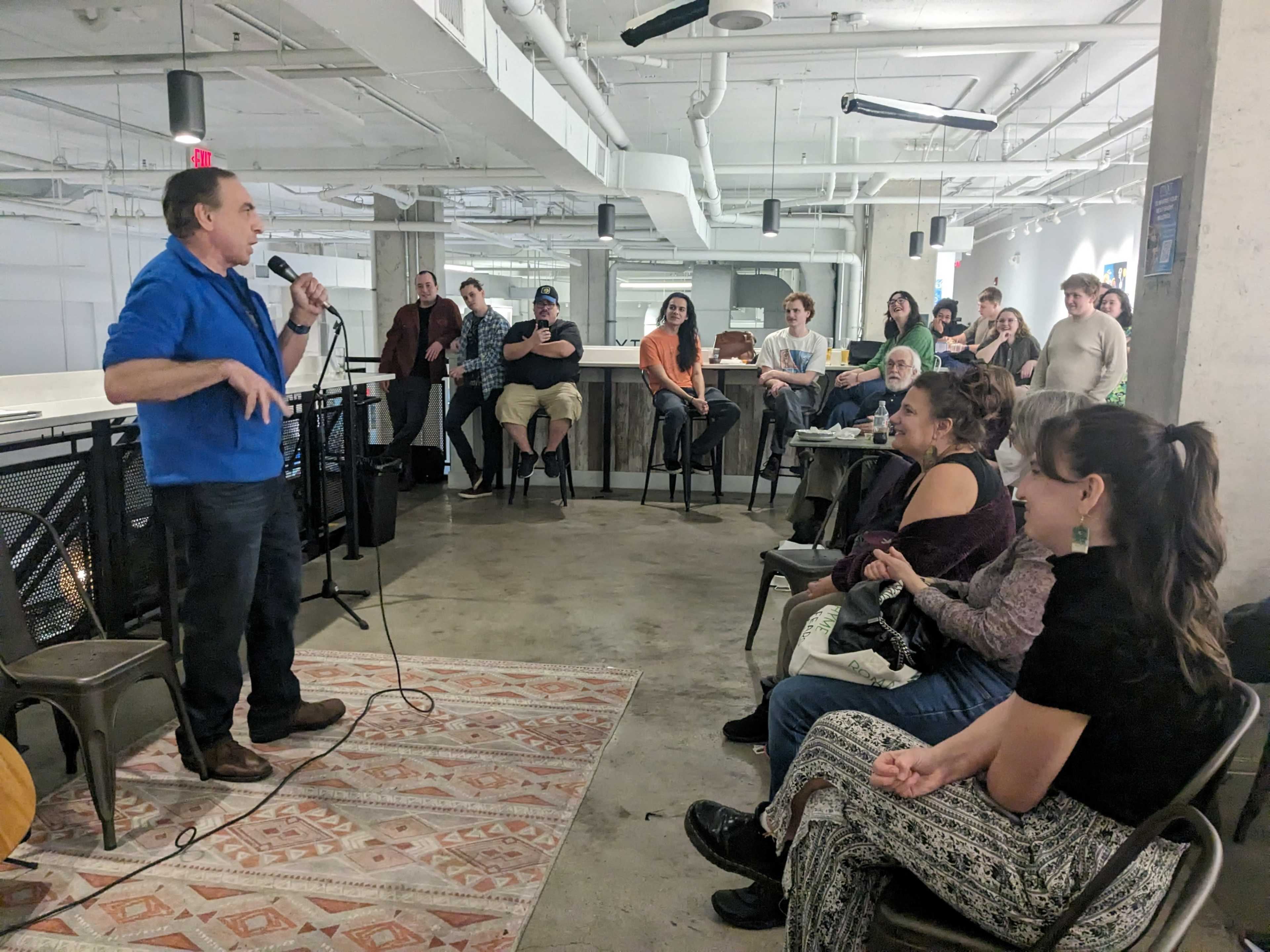 A comedian performs on stage in a casual venue while an audience of various attendees listens and observes from their seats.