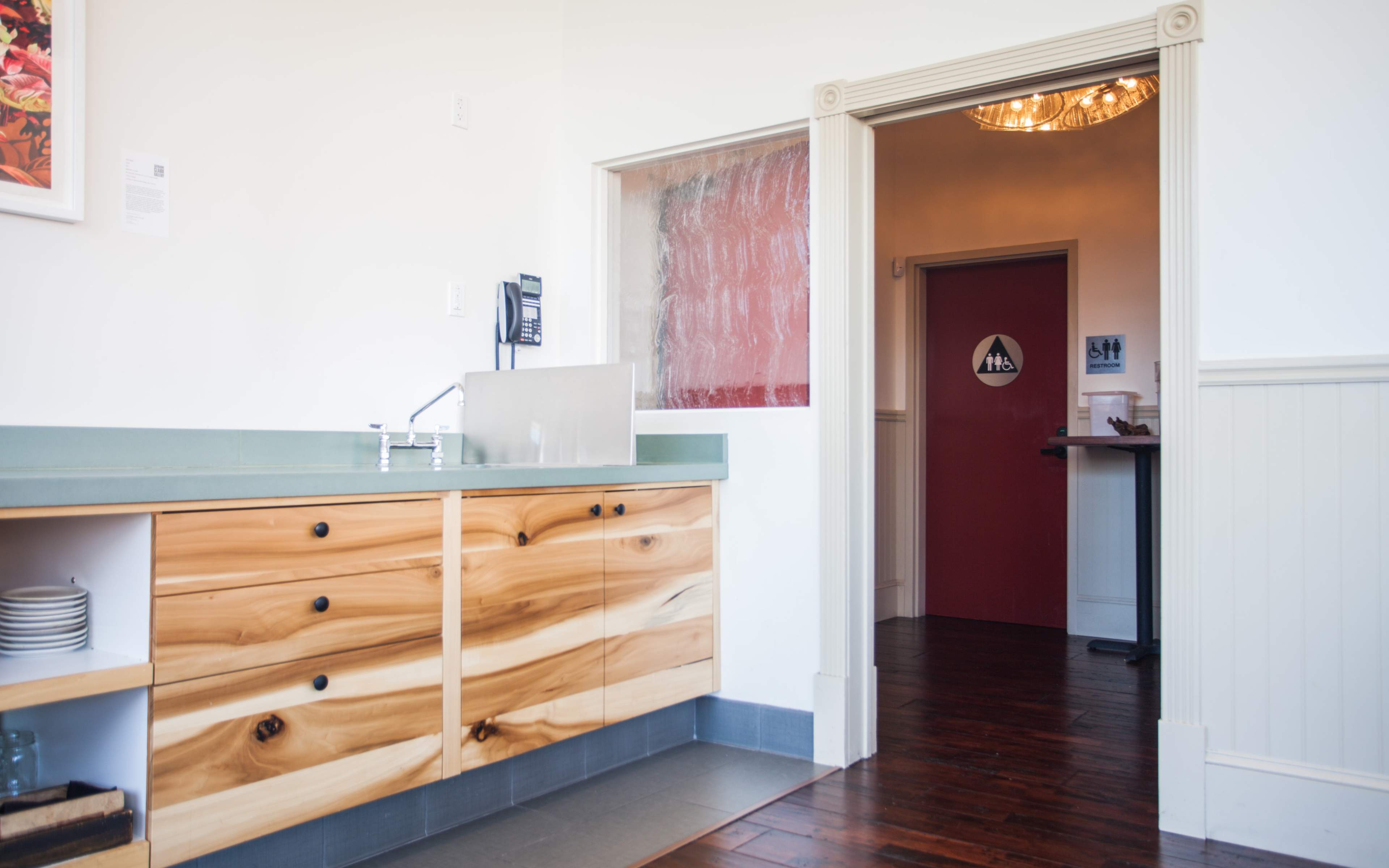 A modern kitchen area with wooden cabinets and a doorway leading to a red-painted room marked with a restroom sign.