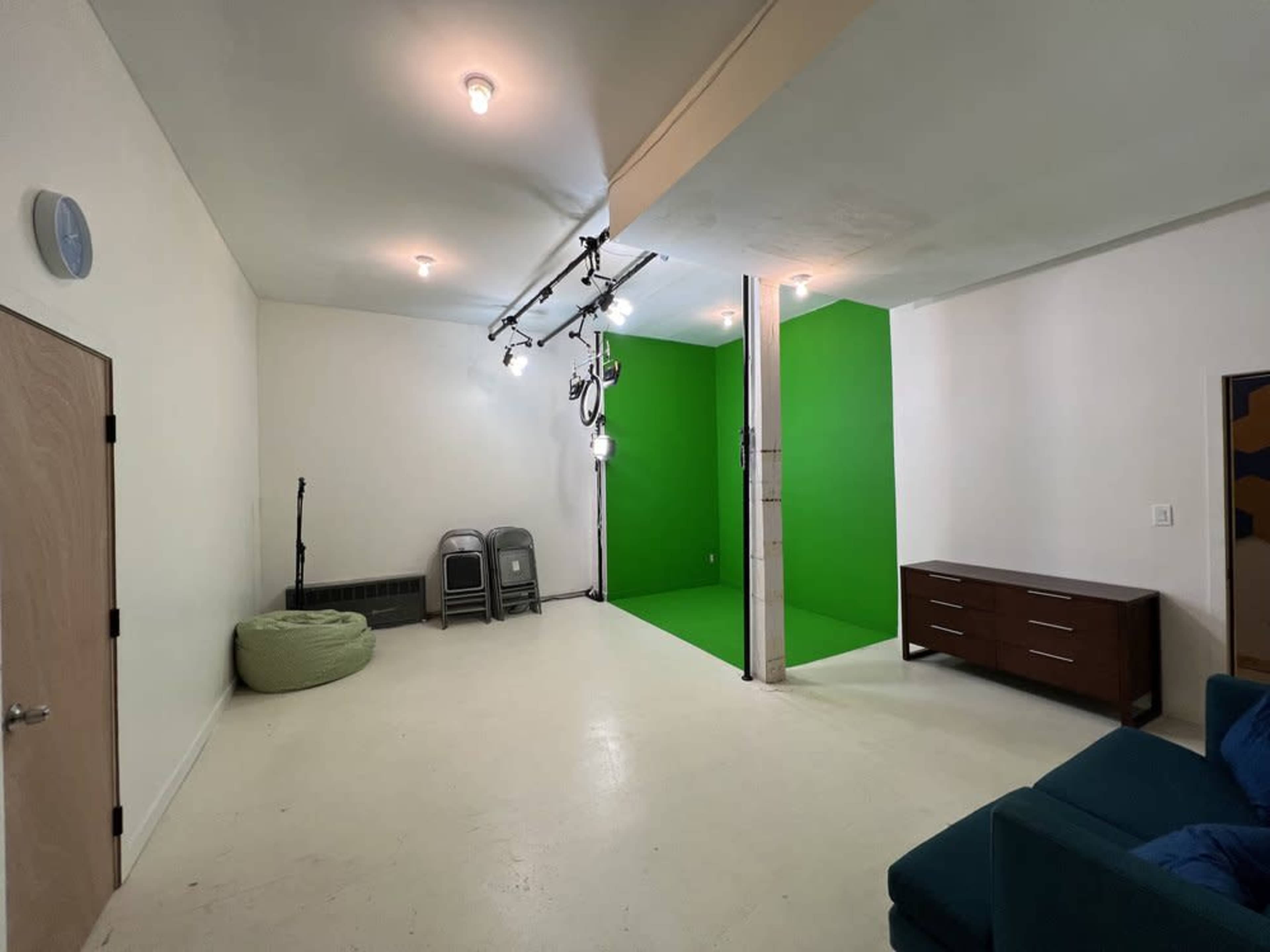 A spacious room featuring a green screen wall, a brown cabinet, two folding chairs, and a green bean bag on a white floor.