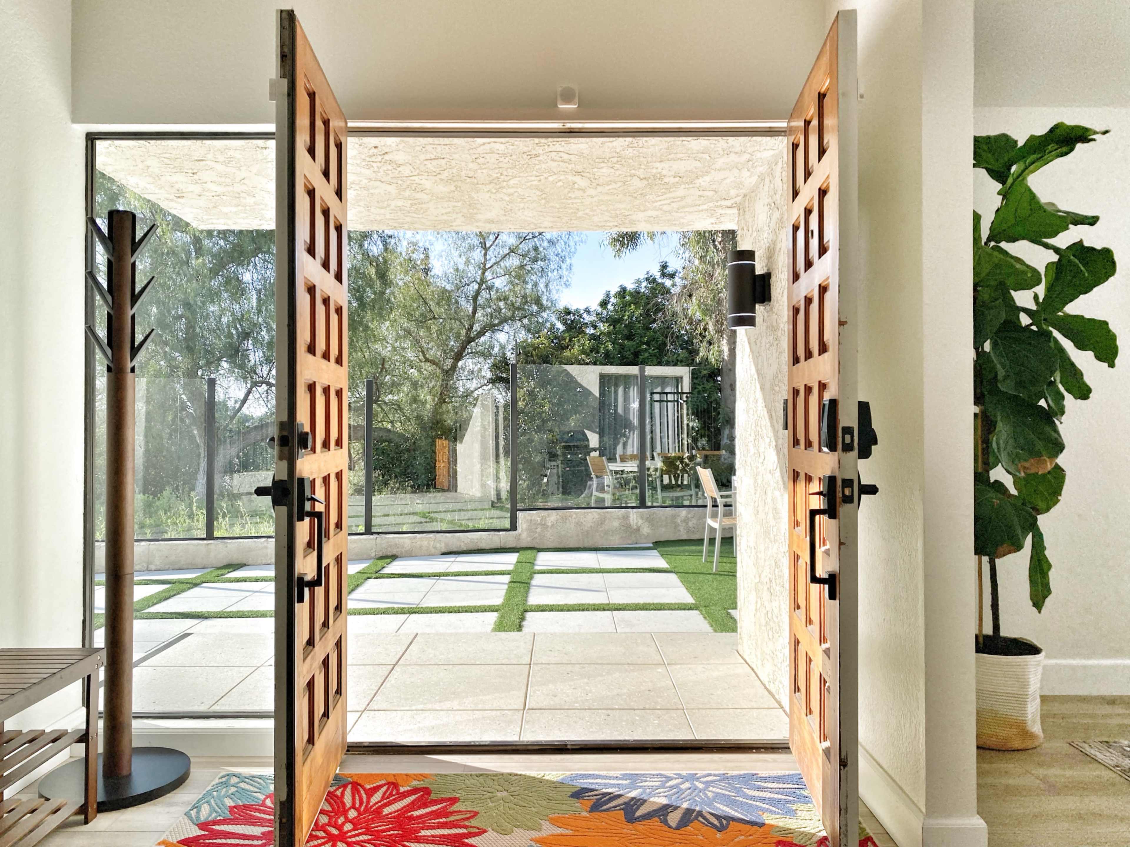 A pair of wooden doors opens to a patio lined with square tiles and greenery, framed by a bright interior setting.