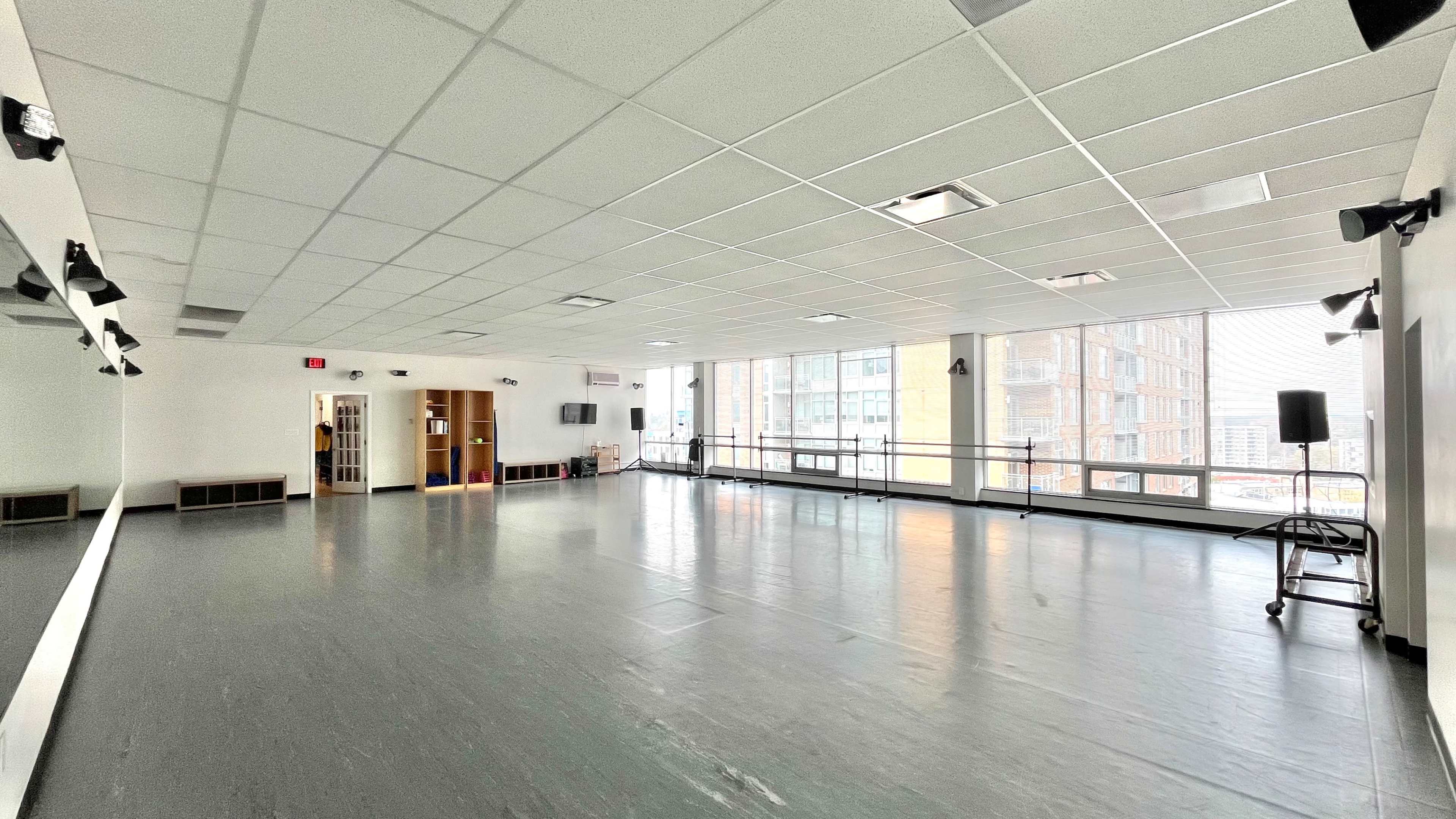 The image shows a spacious dance studio with large windows, a mirrored wall, and polished flooring.