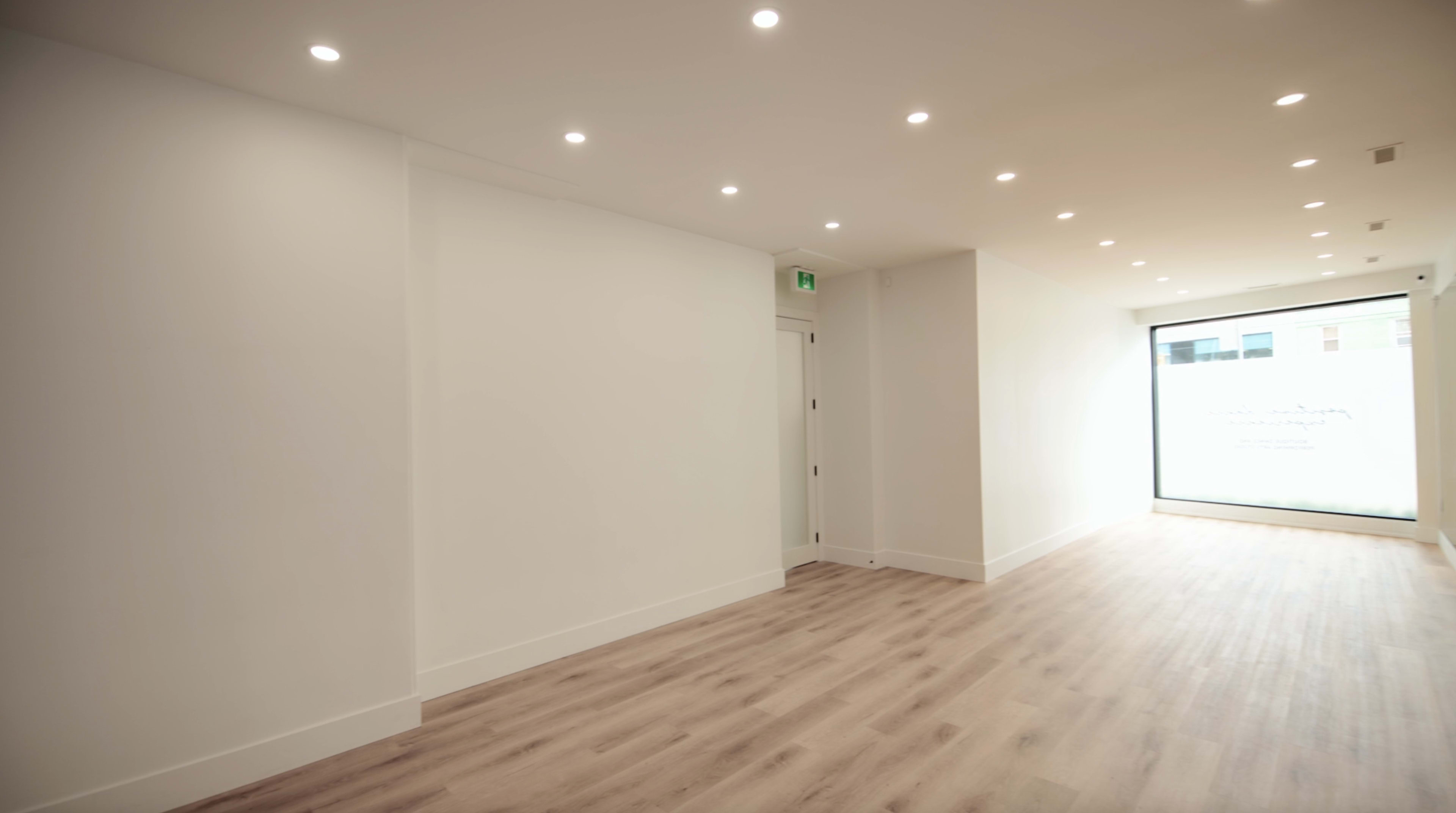 The image shows a bright, empty hallway with light-colored walls and wooden flooring, leading to a large window at the far end.
