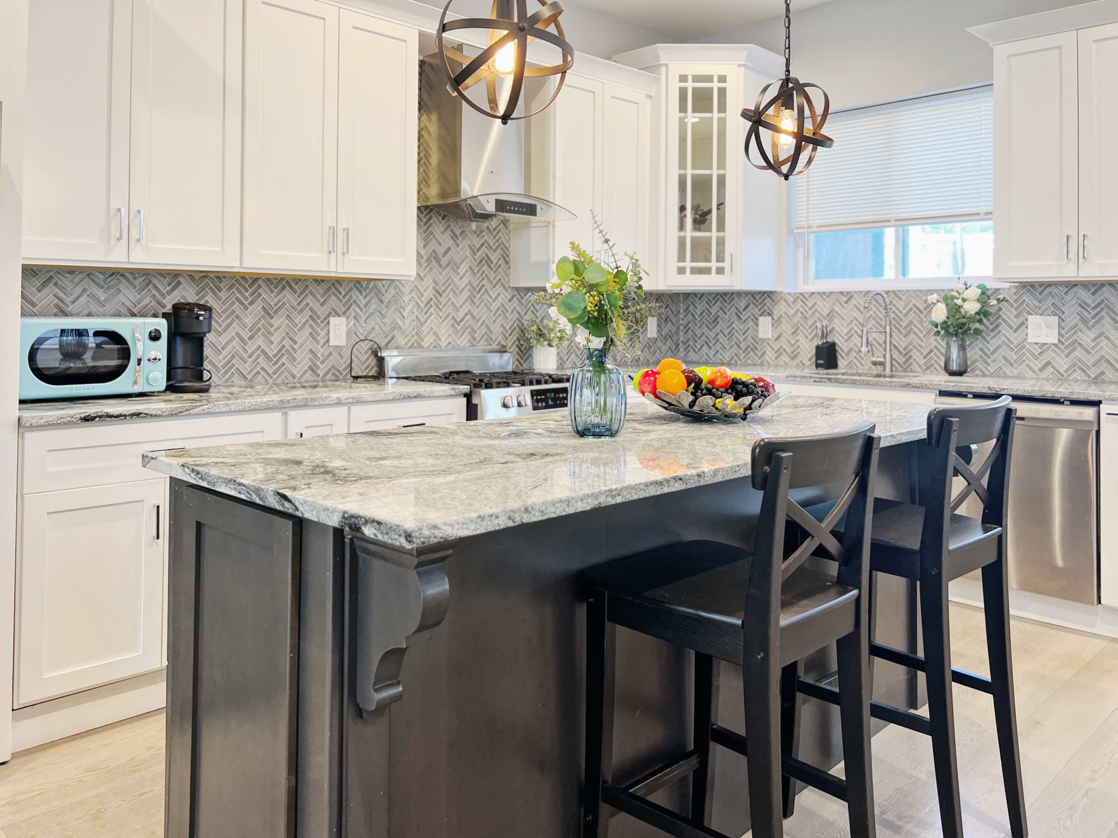 A modern kitchen features white cabinets, a gray marble countertop, and a central island with two black bar stools, surrounded by decorative items and fresh fruit.
