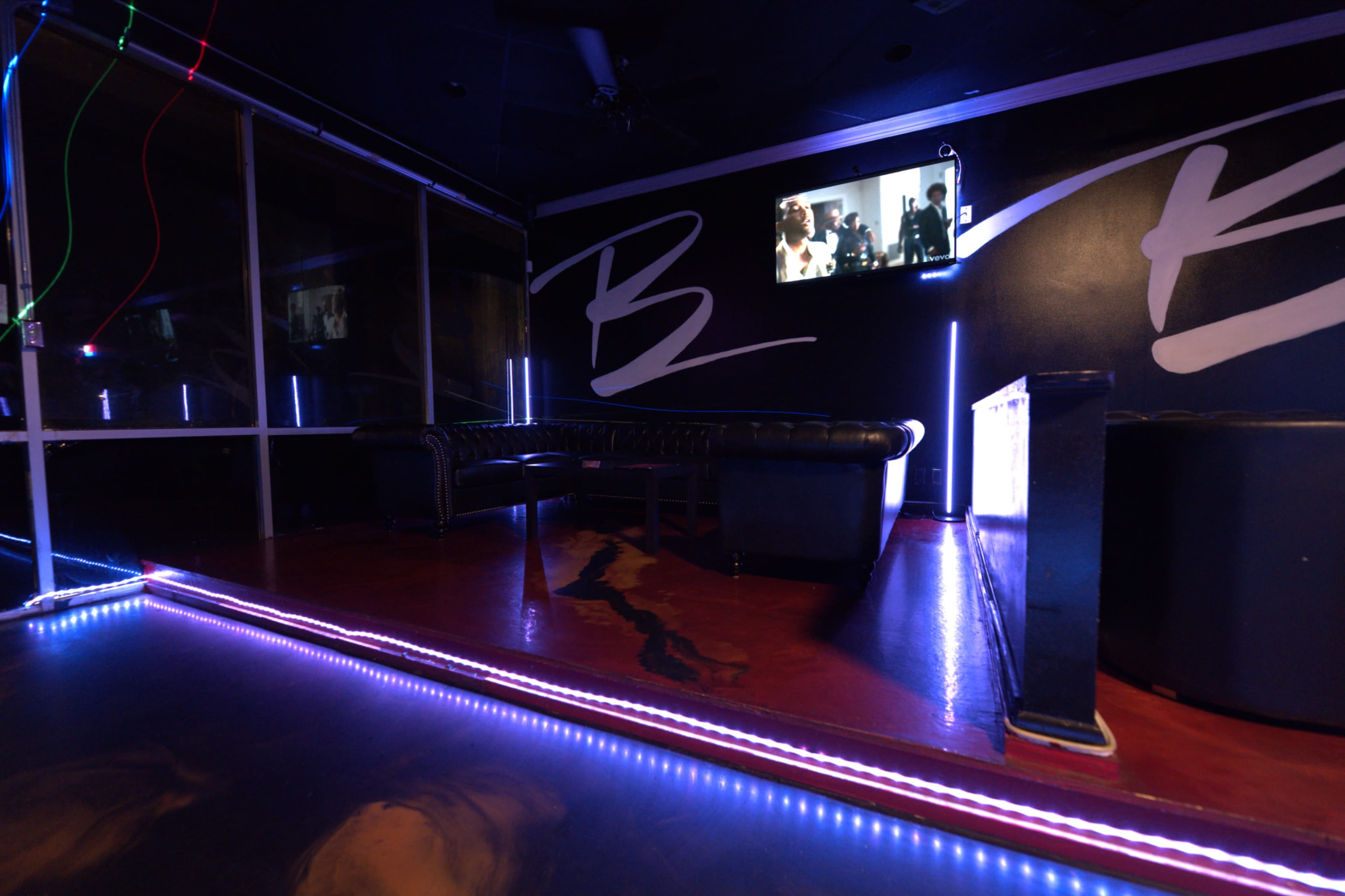 The image depicts a dimly lit lounge area featuring a black leather couch, a bar counter, and a television displaying a scene on the wall.