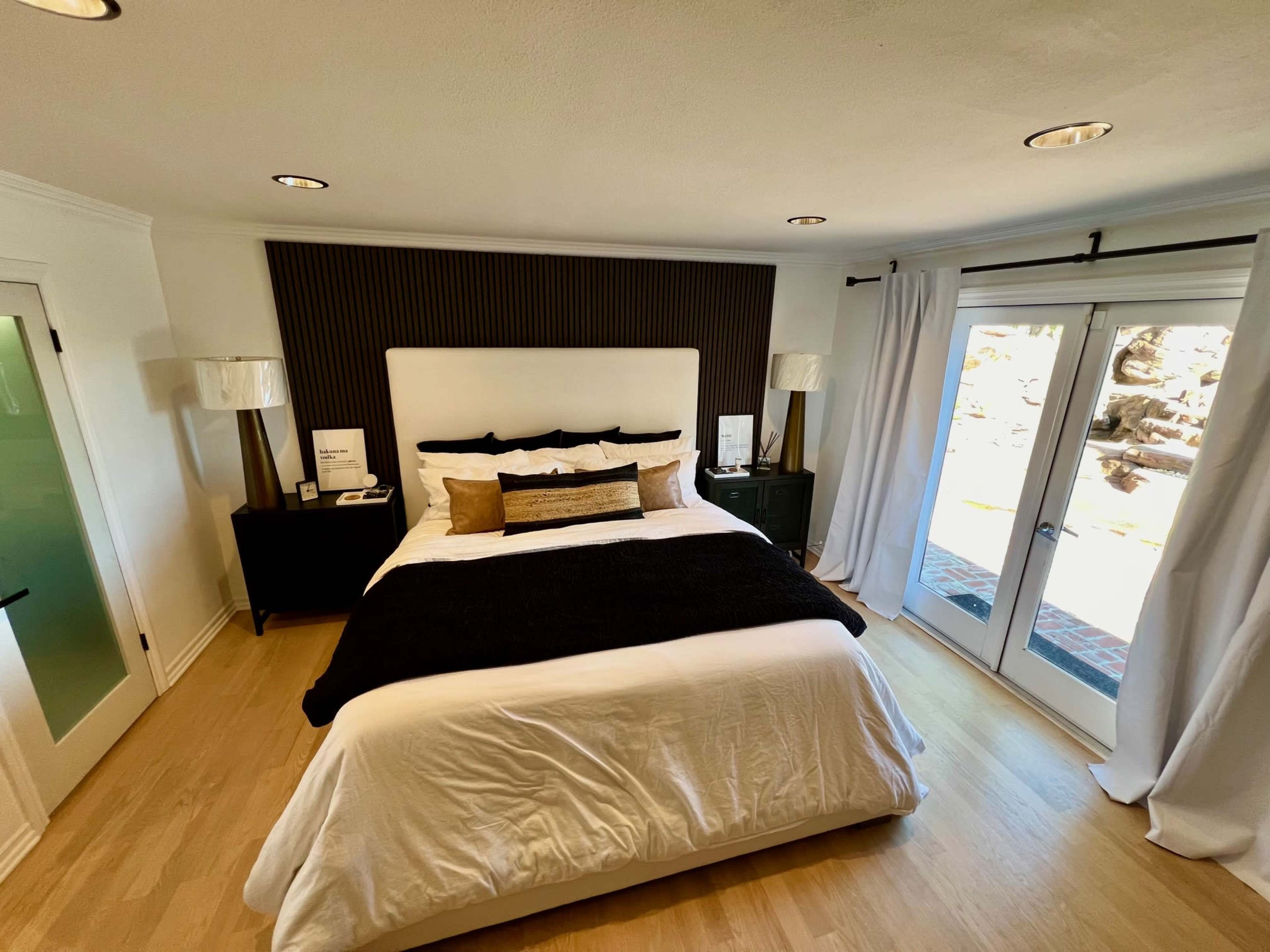 The image shows a modern bedroom with a king-sized bed, a black accent wall, white curtains, and a sliding glass door leading outside.