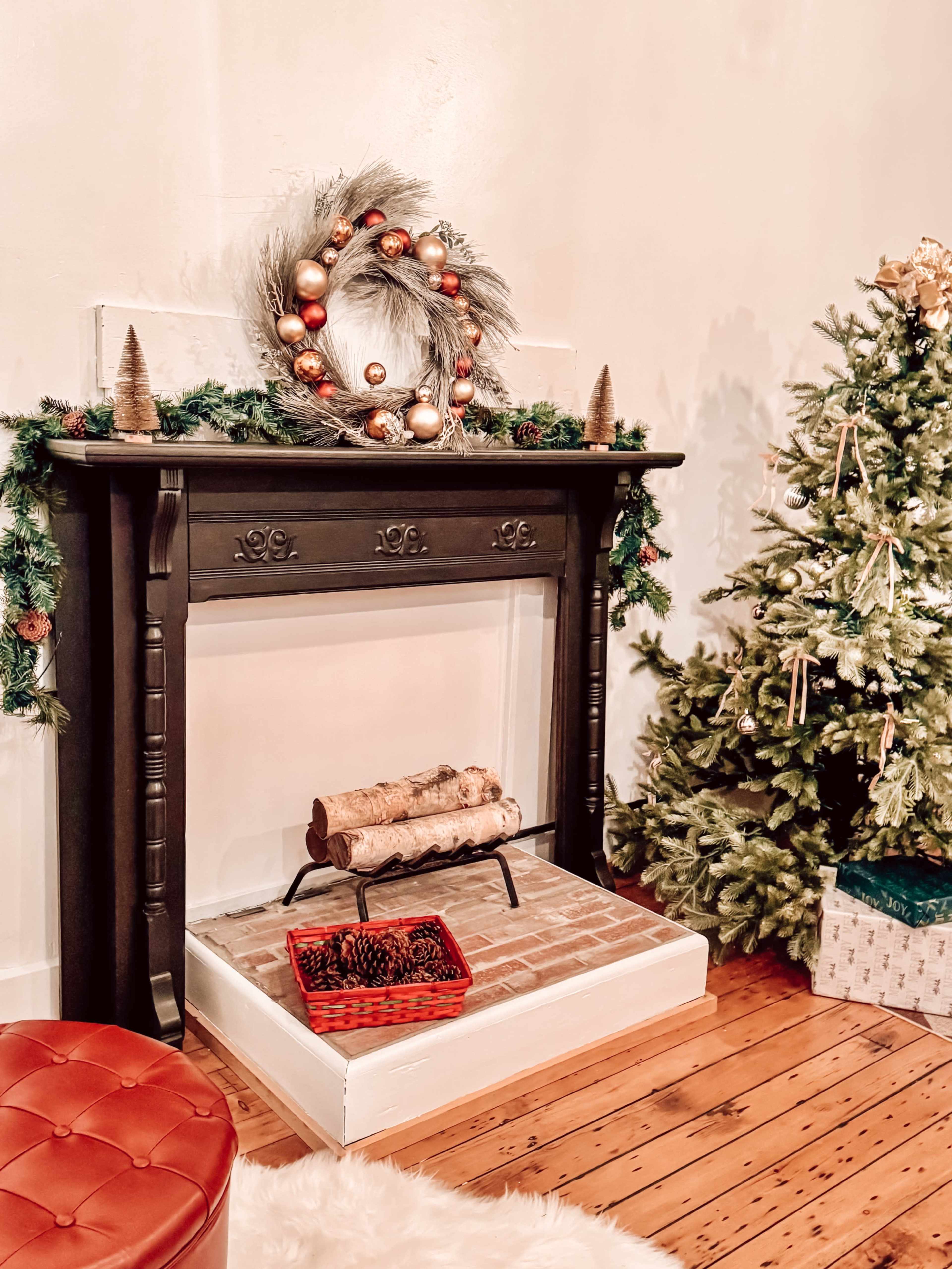 A black fireplace adorned with a wreath and greenery stands beside a decorated Christmas tree in a cozy room.