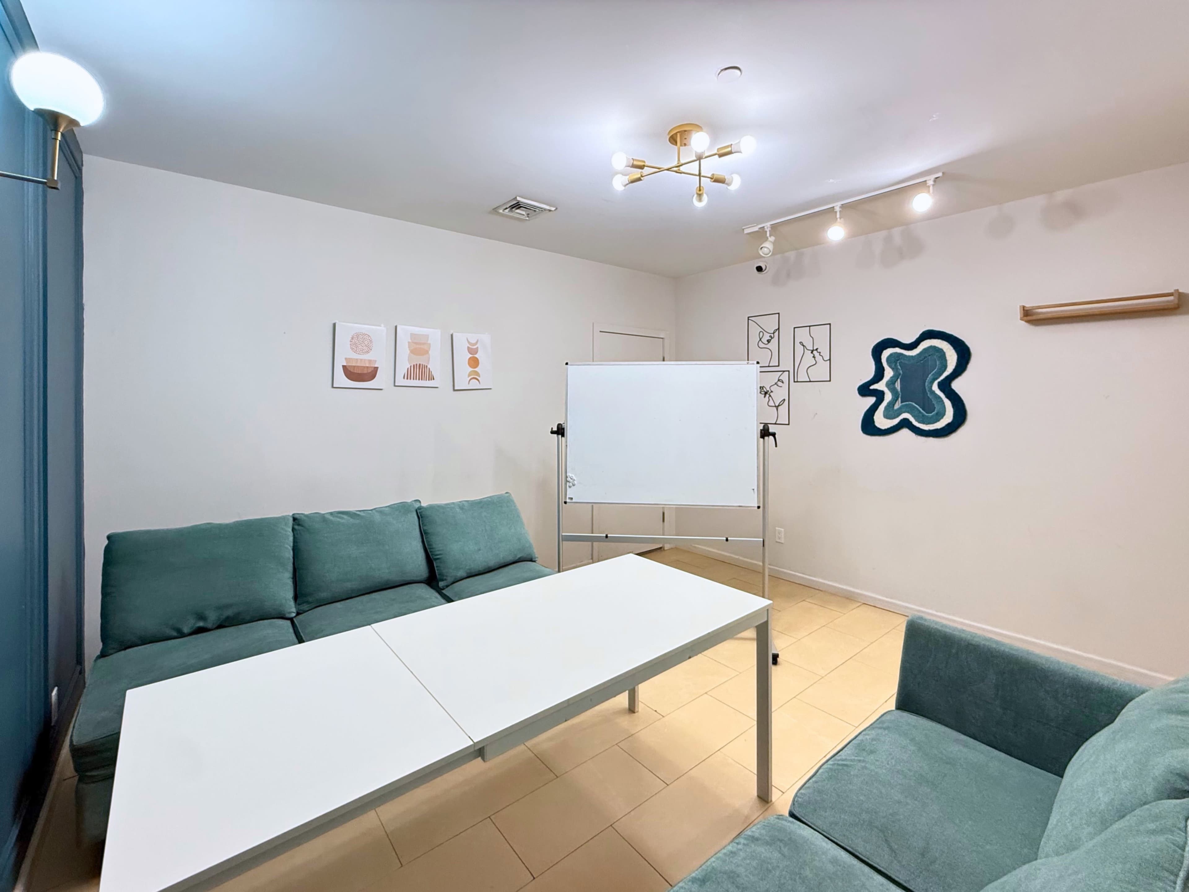 The image shows a small meeting room with two teal sofas, a white table, a flip chart, and framed art on the walls.