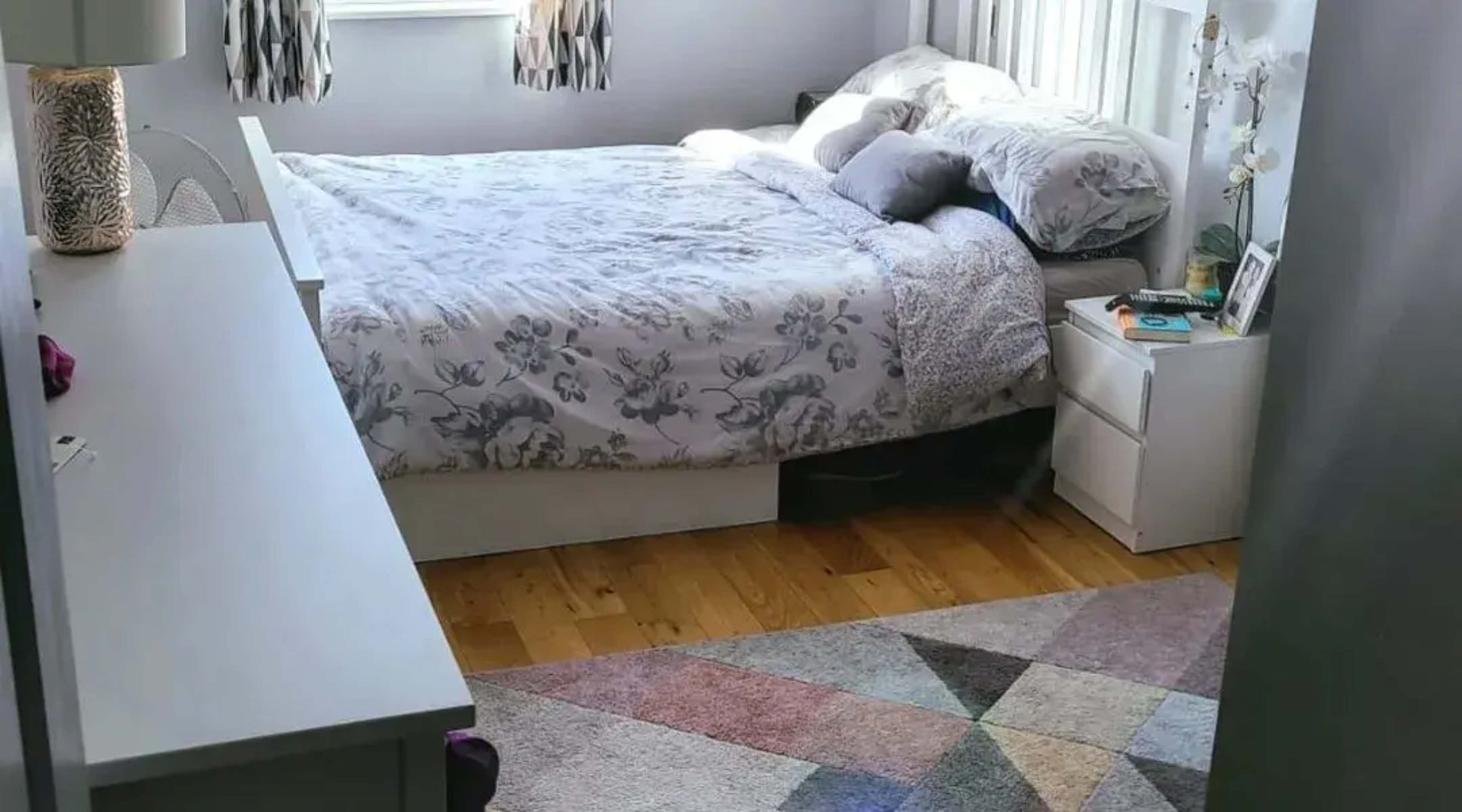 A neatly arranged bedroom featuring a bed with floral bedding, a dresser, a nightstand, and a geometric patterned rug on the wooden floor.