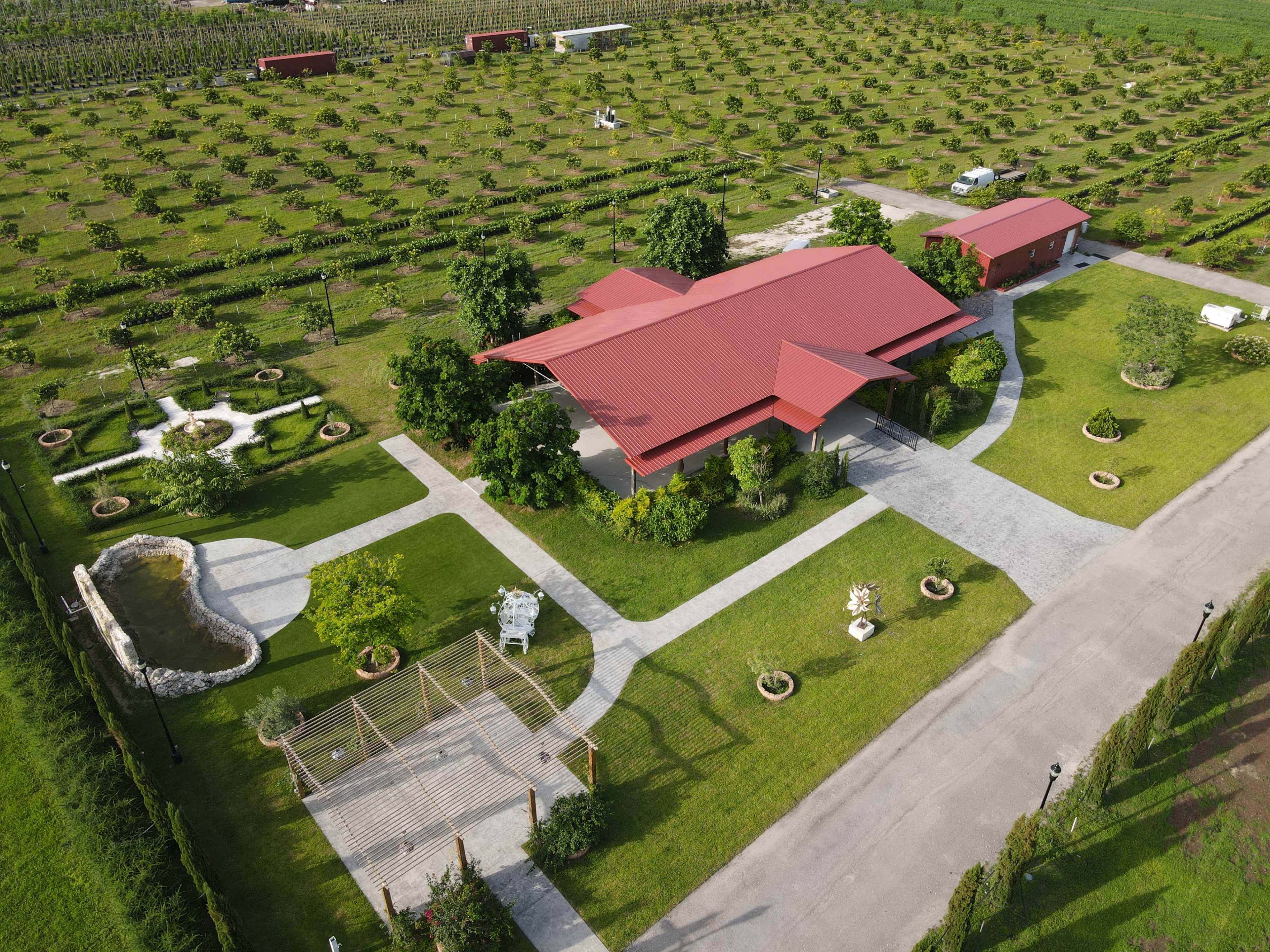 The image shows a modern red-roofed building surrounded by landscaped gardens, pathways, and several trees within an open agricultural area.