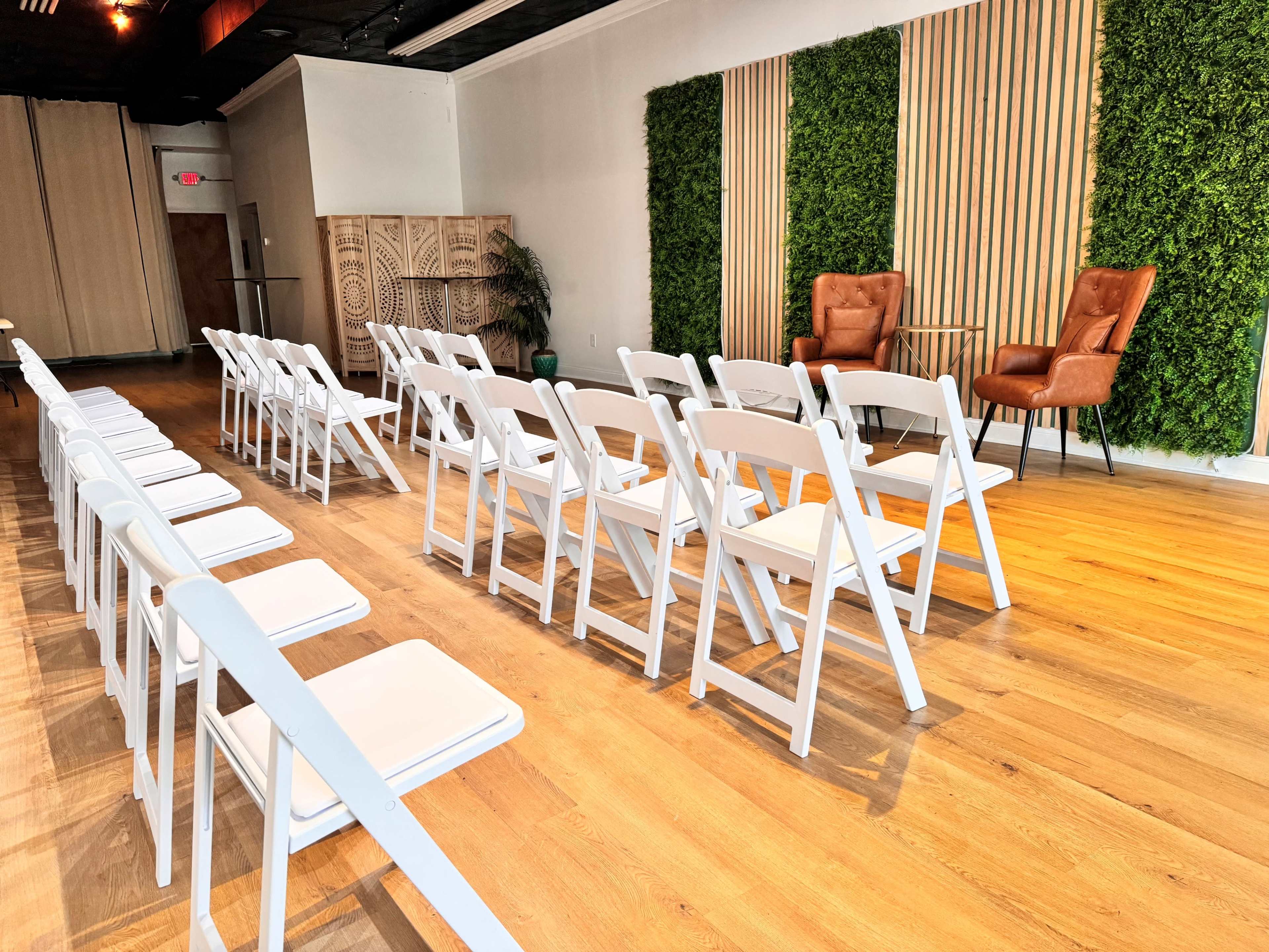 A row of white folding chairs arranged in a spacious room with a green wall and two brown armchairs set against a wooden backdrop.