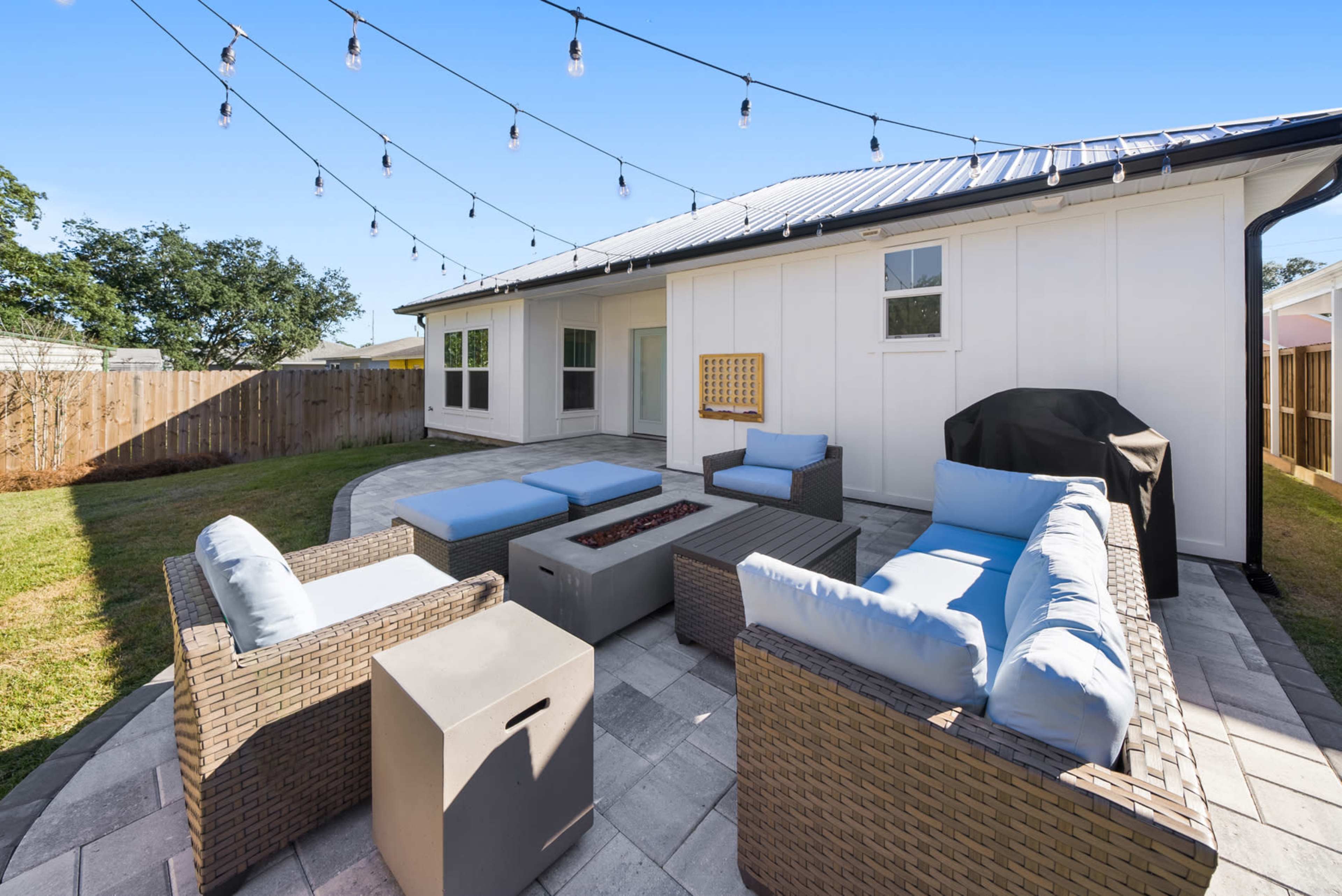A backyard patio area with seating arrangements, a fire pit, and string lights above, surrounded by grass and a wooden fence.