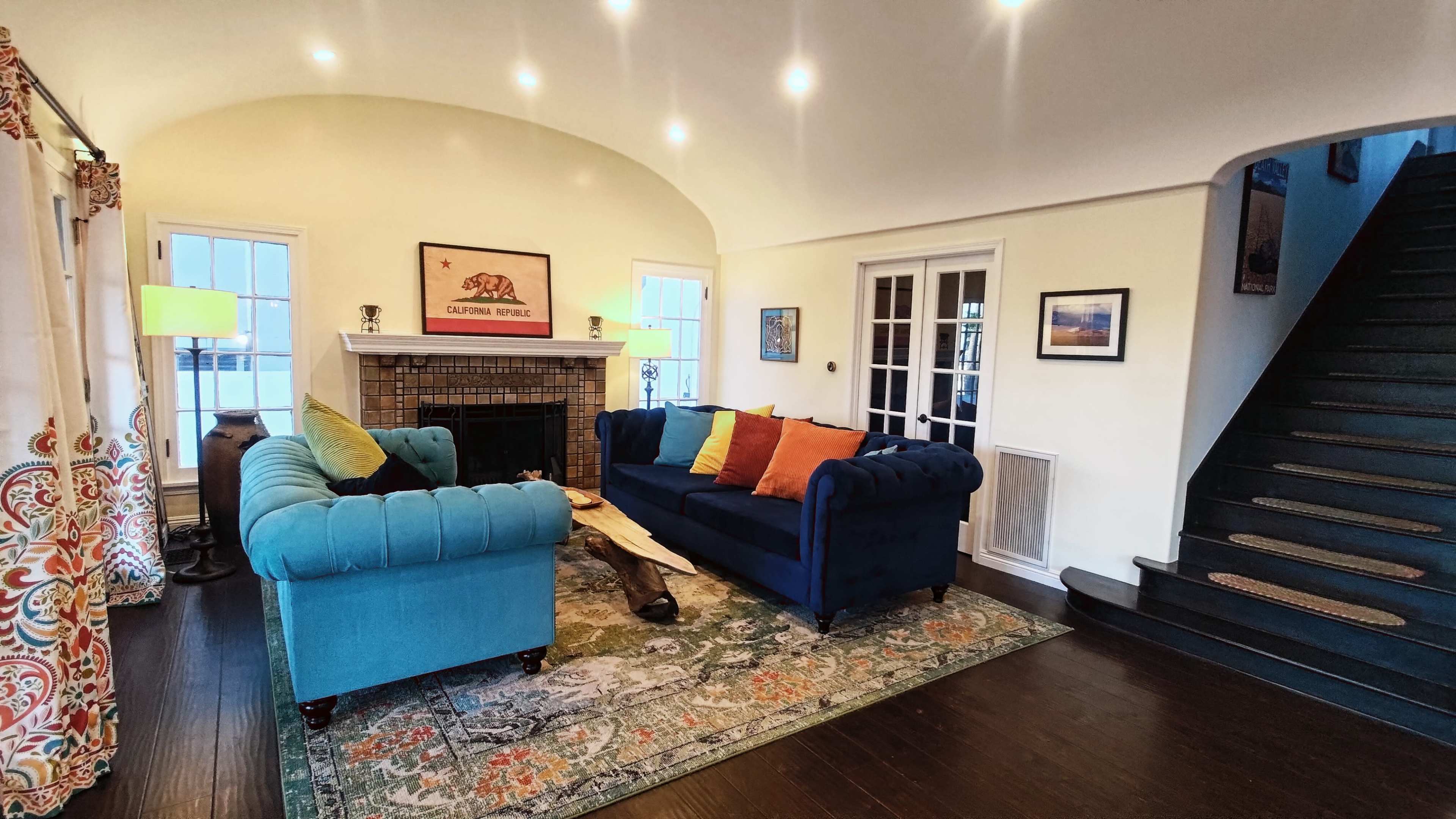 The image shows a cozy living room featuring a blue sofa with colorful pillows, a fireplace, a decorative rug, and a staircase in the background.
