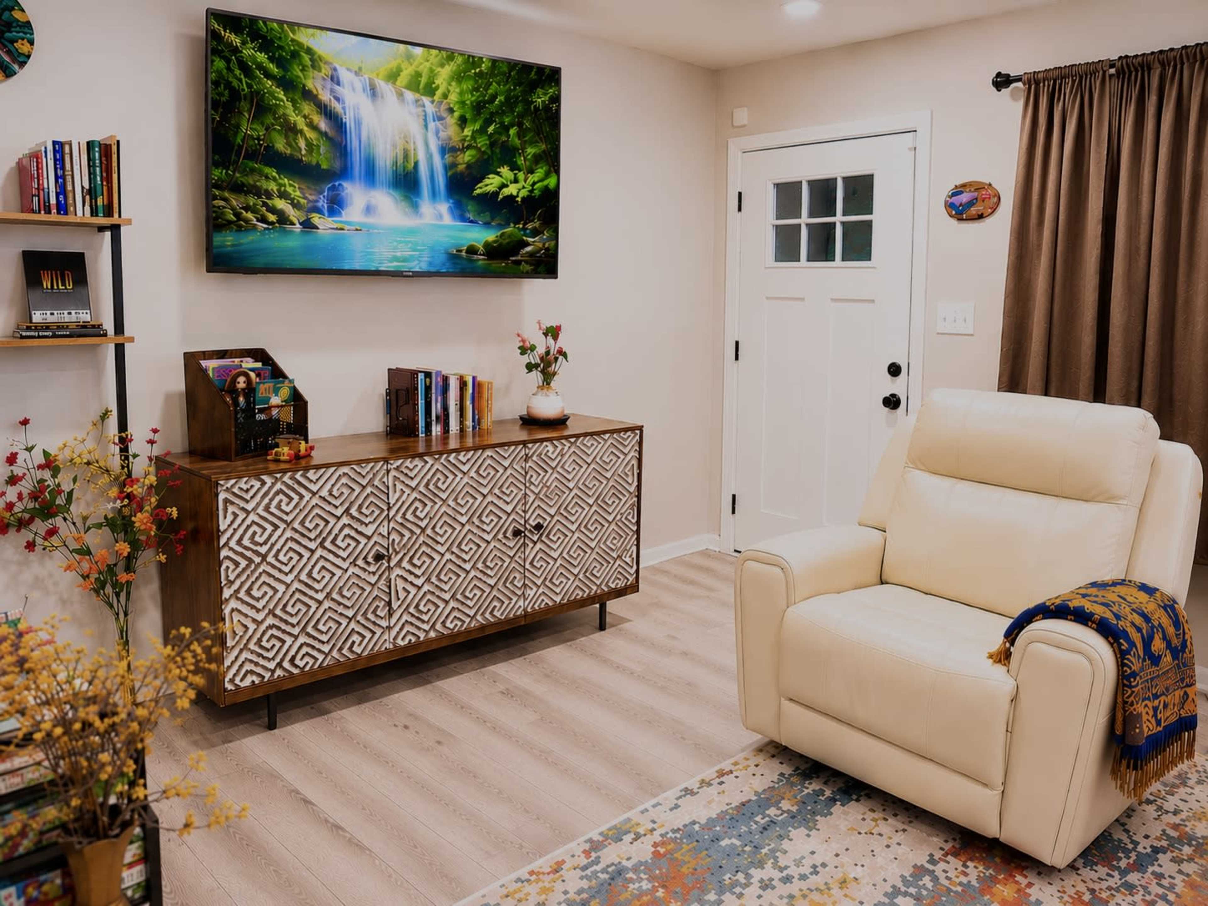The image shows a cozy living room featuring a white reclining chair, a wooden TV stand with decorative patterns, a television displaying a waterfall scene, and a door opposite the chair.