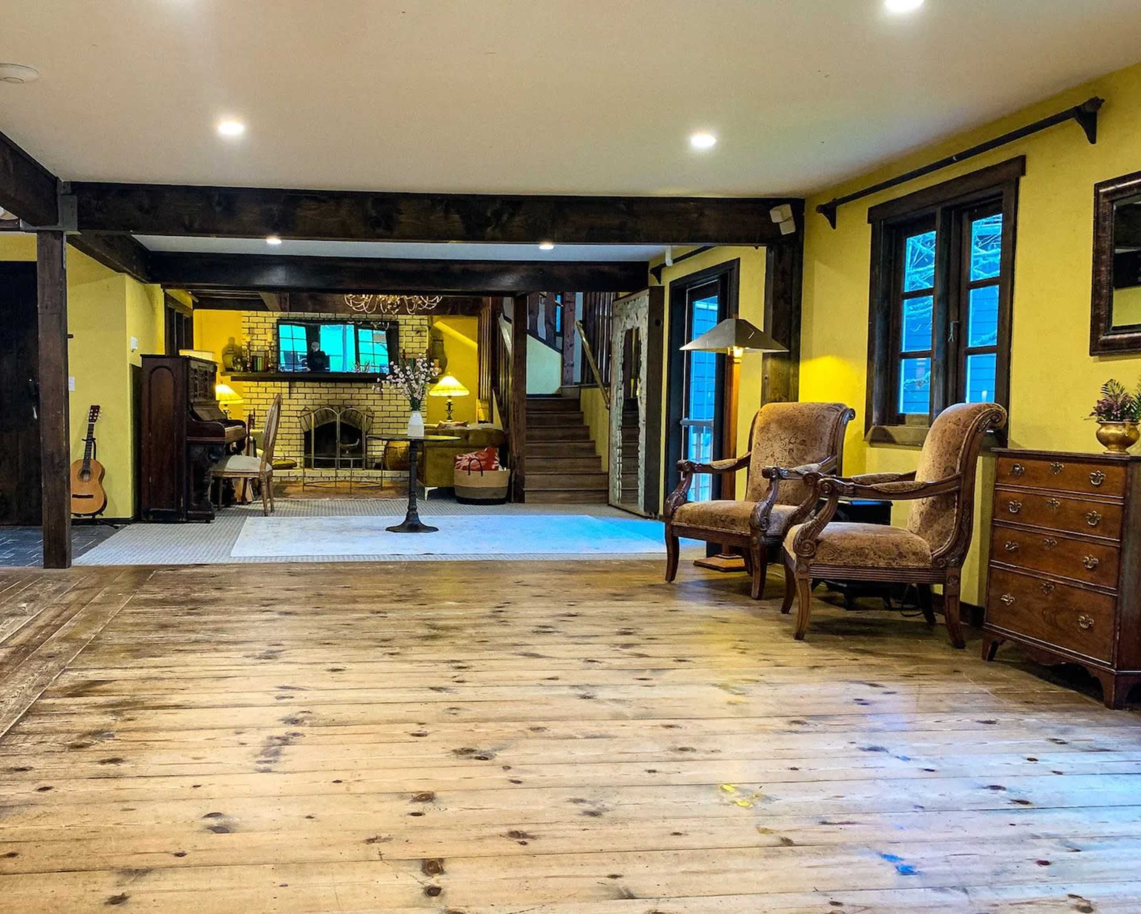 The image shows a spacious interior of a home with wooden floors, yellow walls, and a staircase leading to a second level, furnished with two armchairs, a cabinet, and a piano.