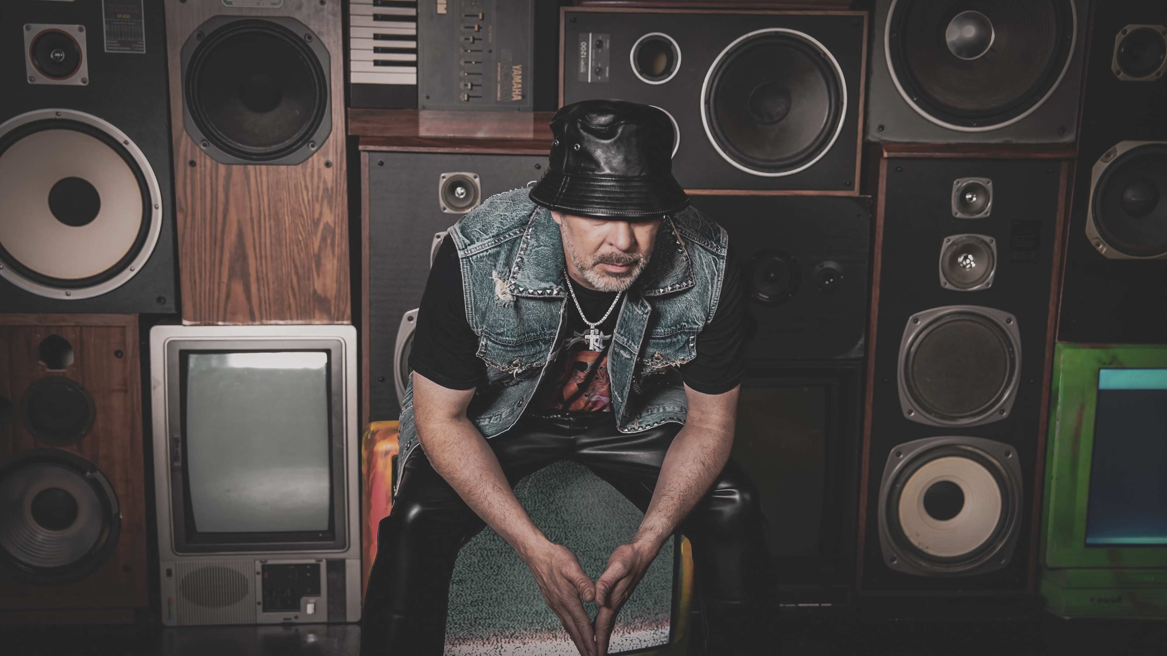 A man wearing a leather bucket hat and a denim vest sits on a chair in front of a wall of speakers and old television sets.