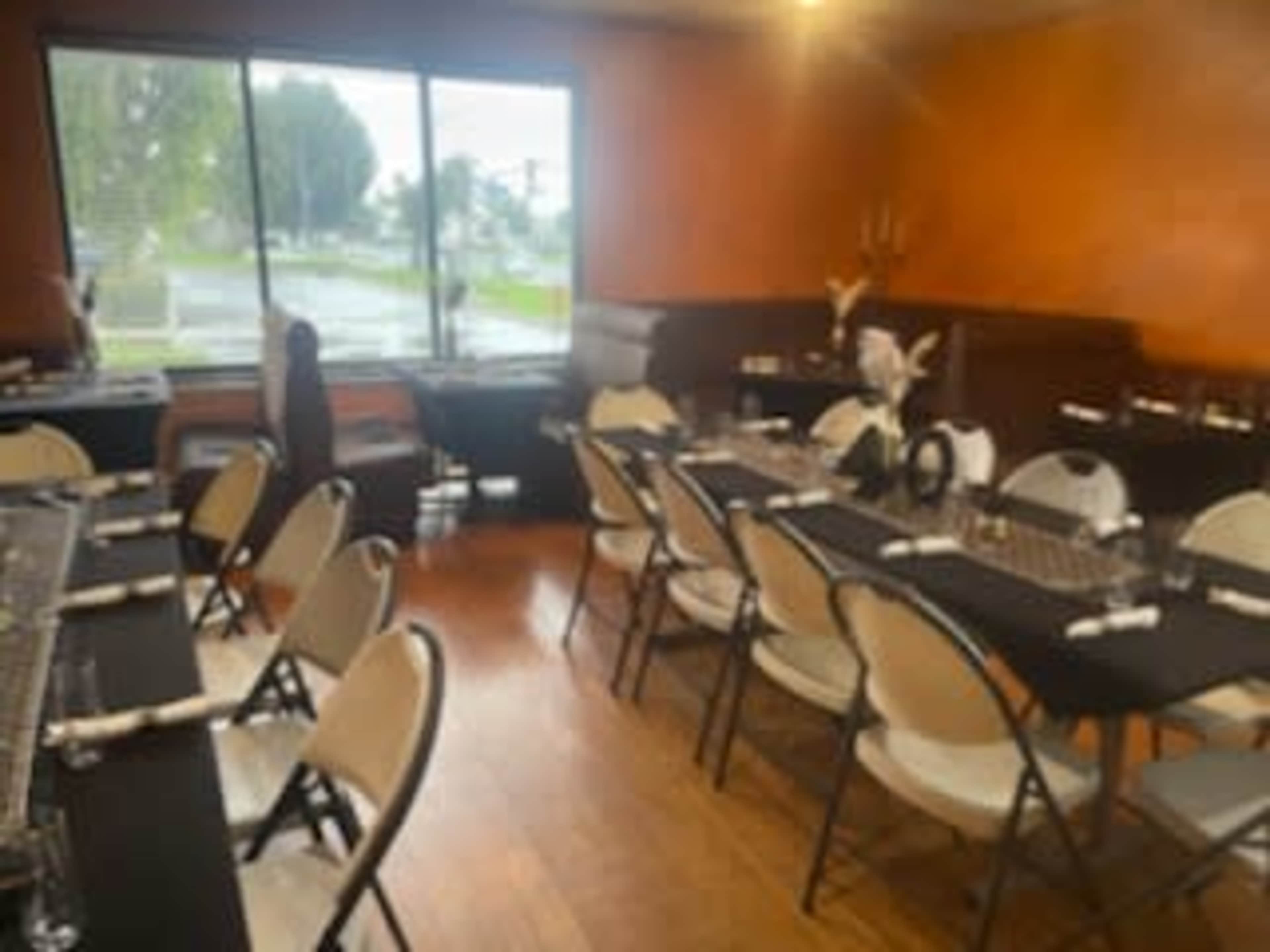 A dining area is set with several tables and chairs, featuring black tablecloths and white napkins, with large windows showing a rainy outdoor scene.