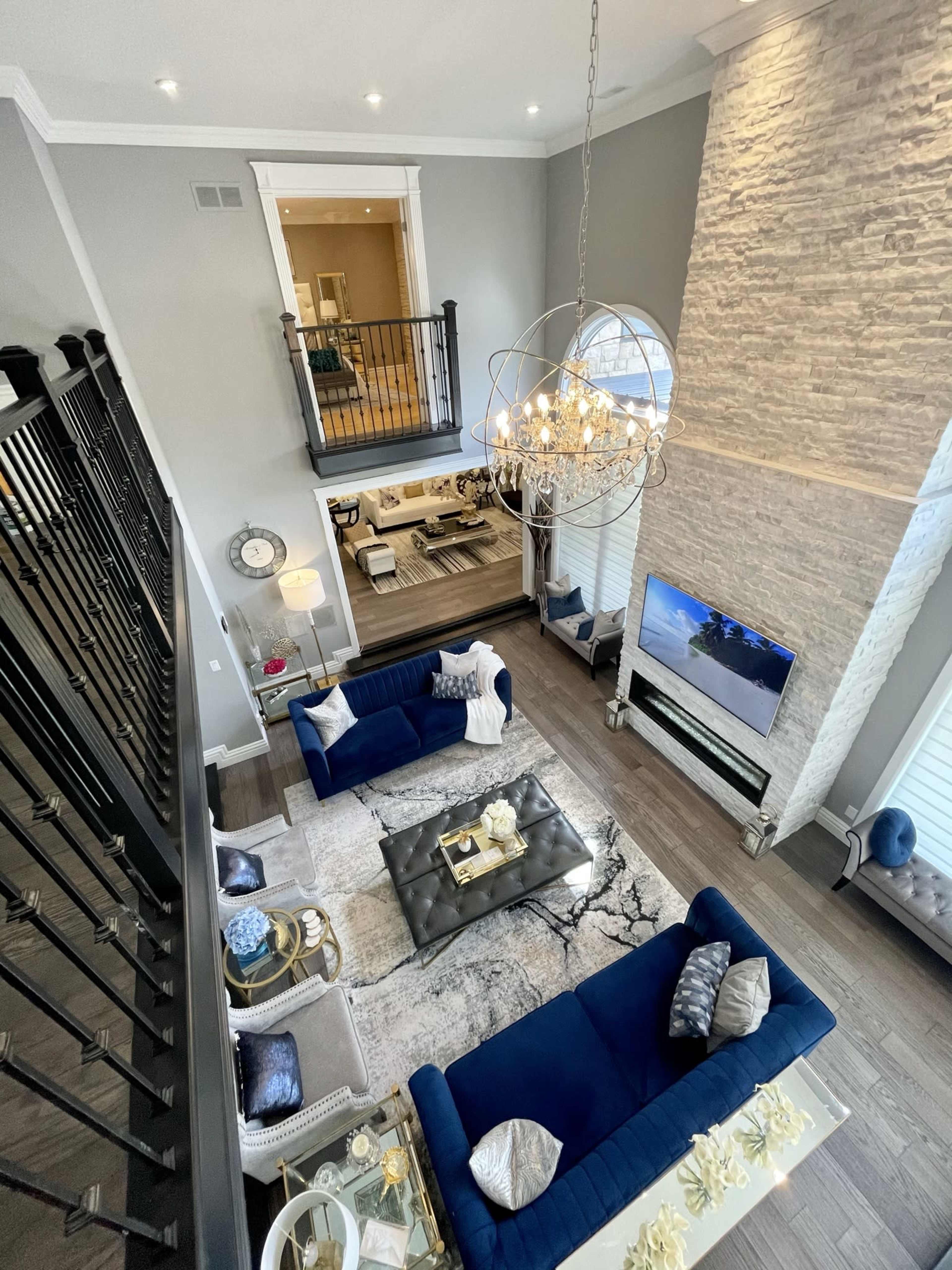 An elegant two-story living room with navy blue sofas, a large chandelier, and a stone accent wall, featuring a TV mounted above a modern fireplace.