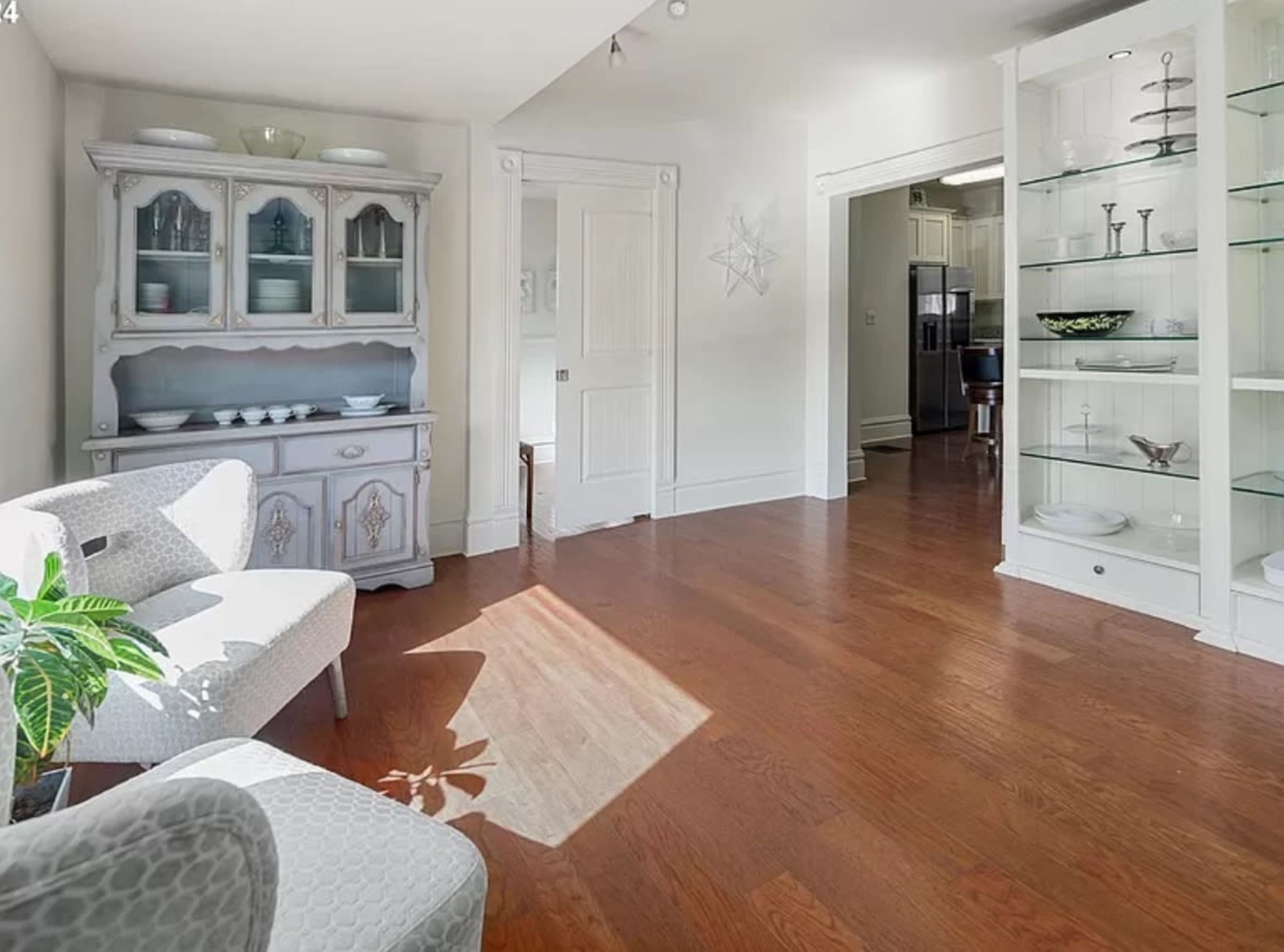A brightly lit living space featuring a light-colored cabinet and a gray armchair, with wooden flooring and a doorway leading to a kitchen area.