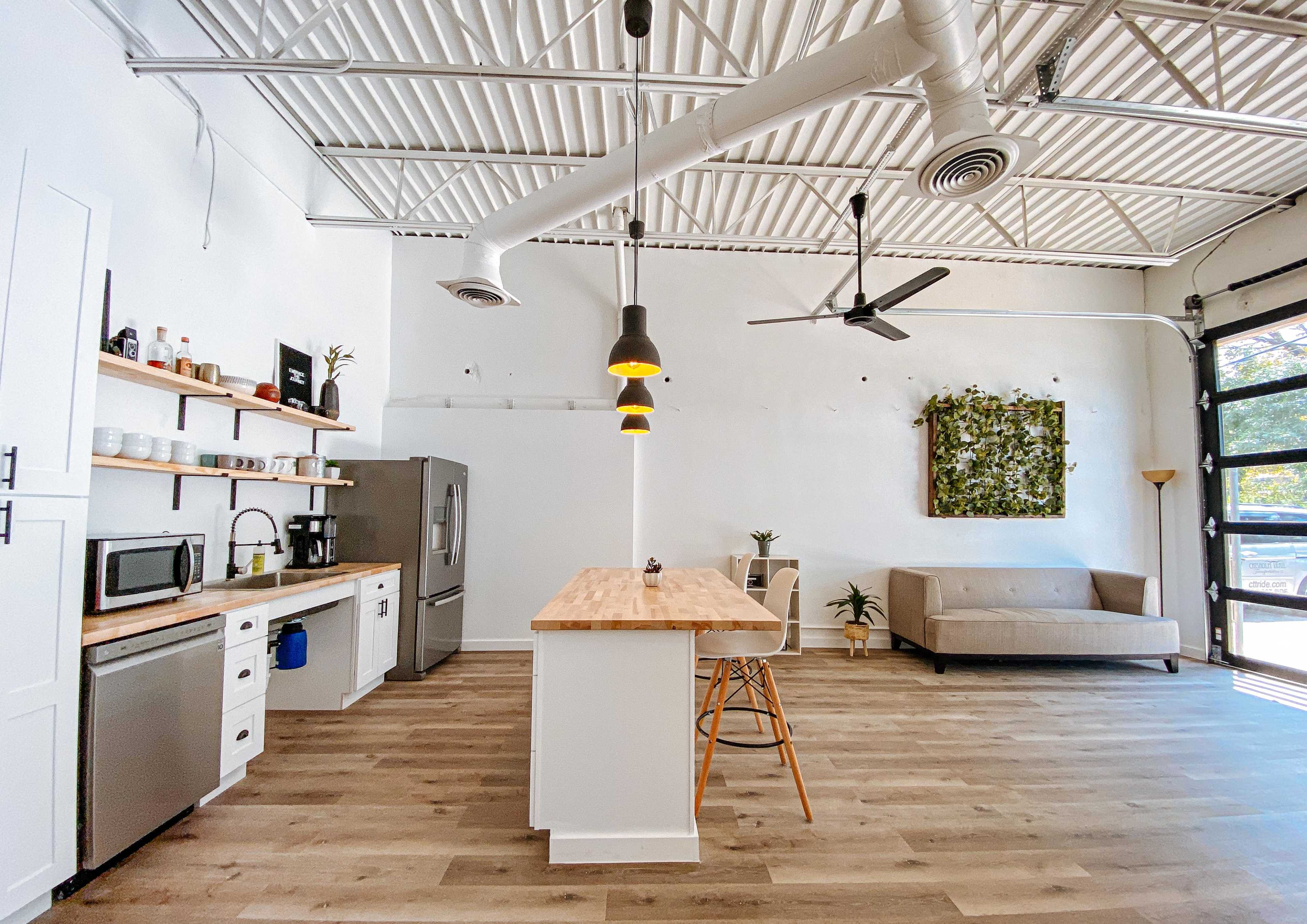 A bright and spacious kitchen area features a wooden island with bar stools, a stainless steel refrigerator, and a couch against the wall, complemented by green plants and a wall-mounted artwork.