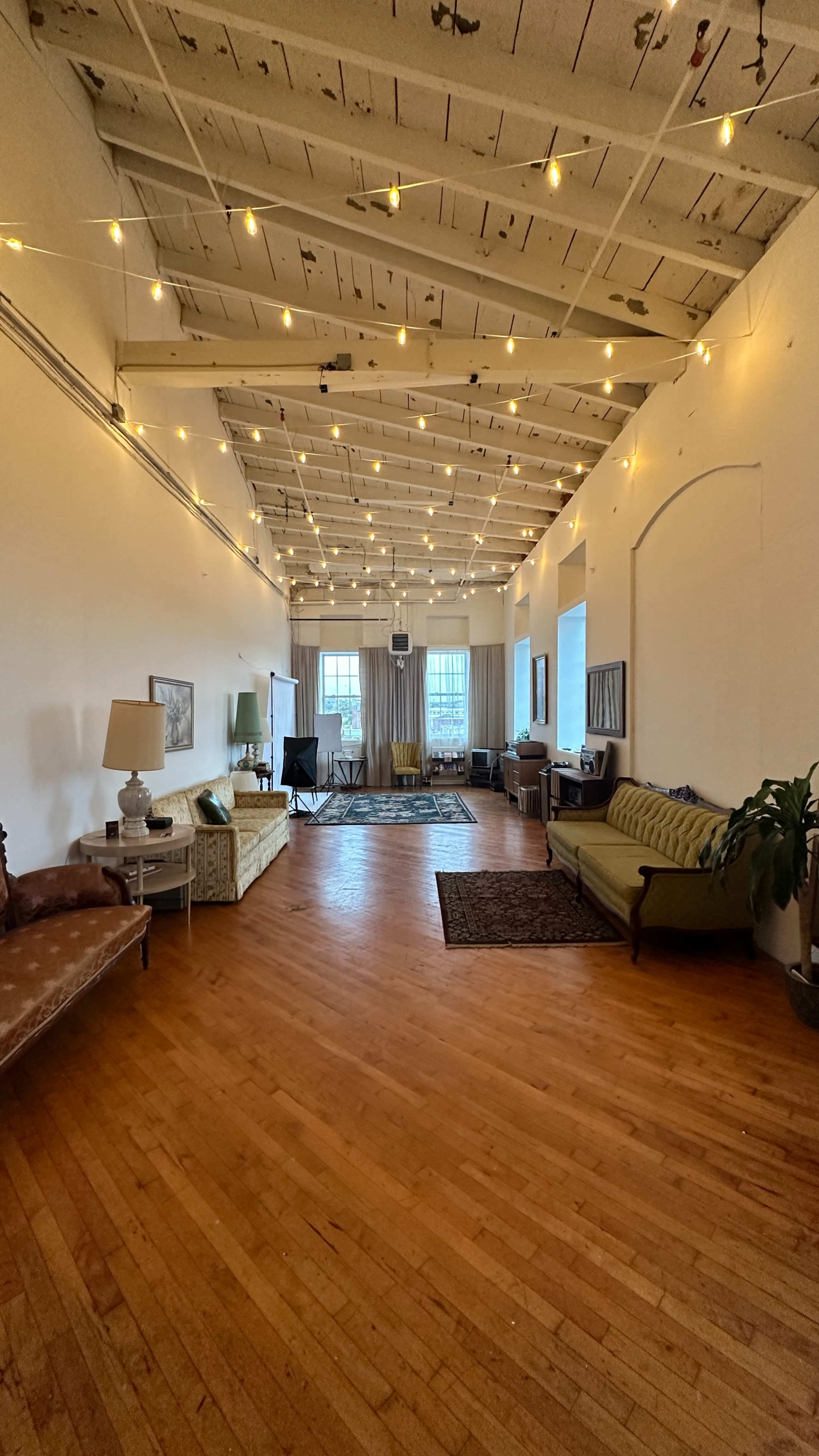 A spacious, well-lit room with wooden flooring, string lights hanging from the ceiling, and a mix of vintage furniture along the walls.