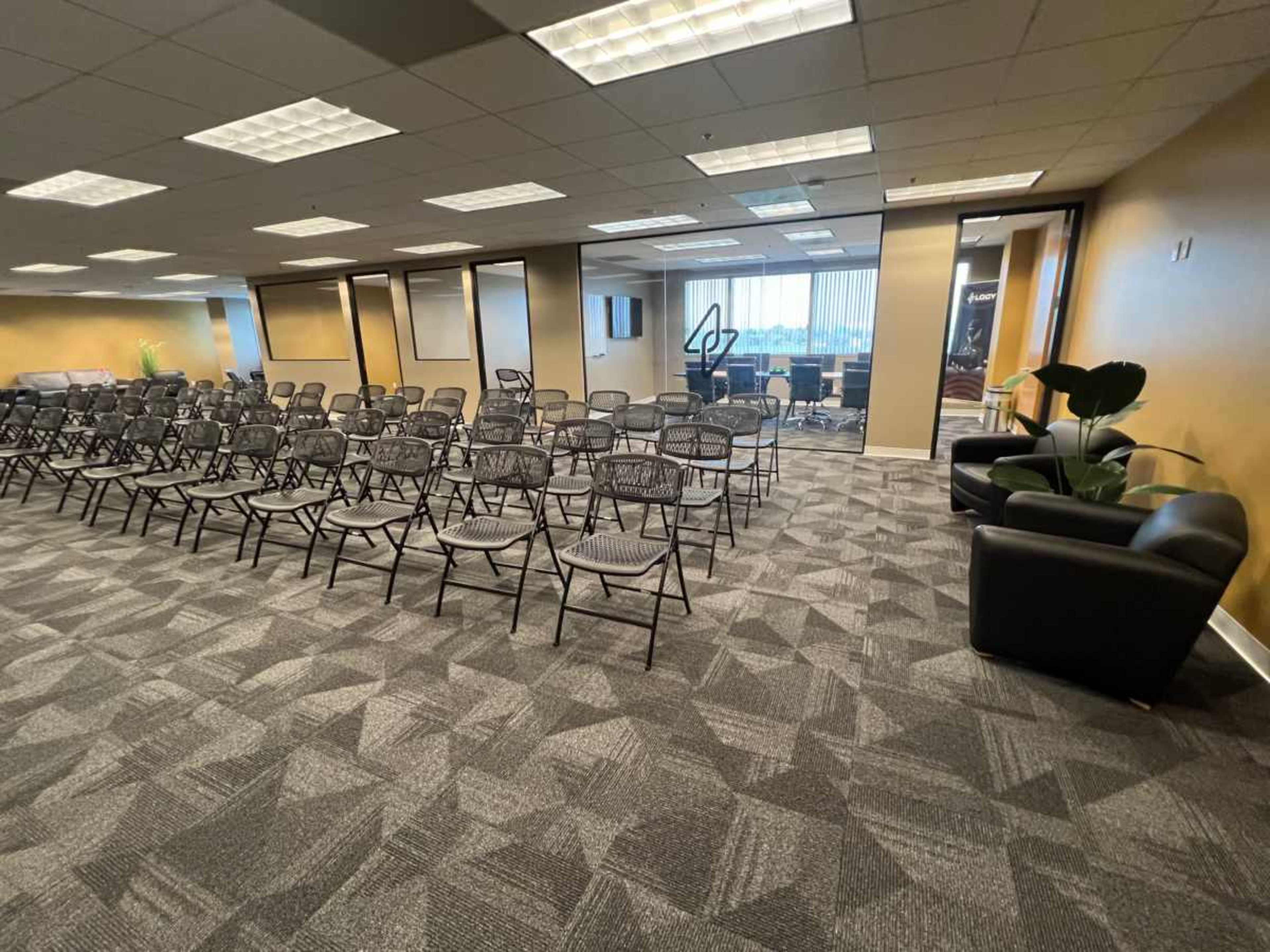 The image shows a spacious office meeting room arranged with rows of chairs and a seating area featuring a couch and a plant.
