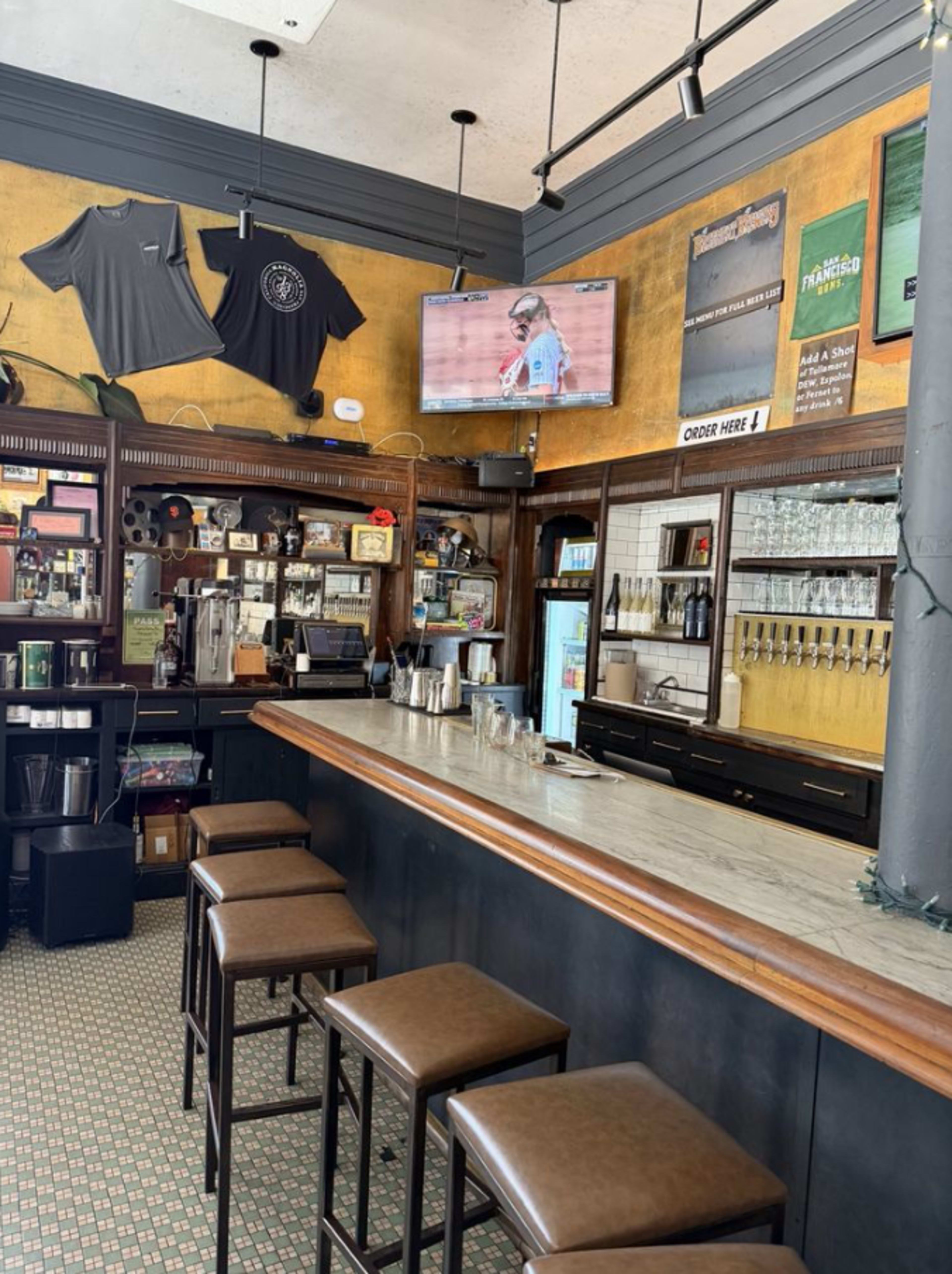 The image shows a bar interior with a marble countertop, high stools, and a television displaying a sports event, surrounded by shelves stocked with drinks and various decorations on the walls.