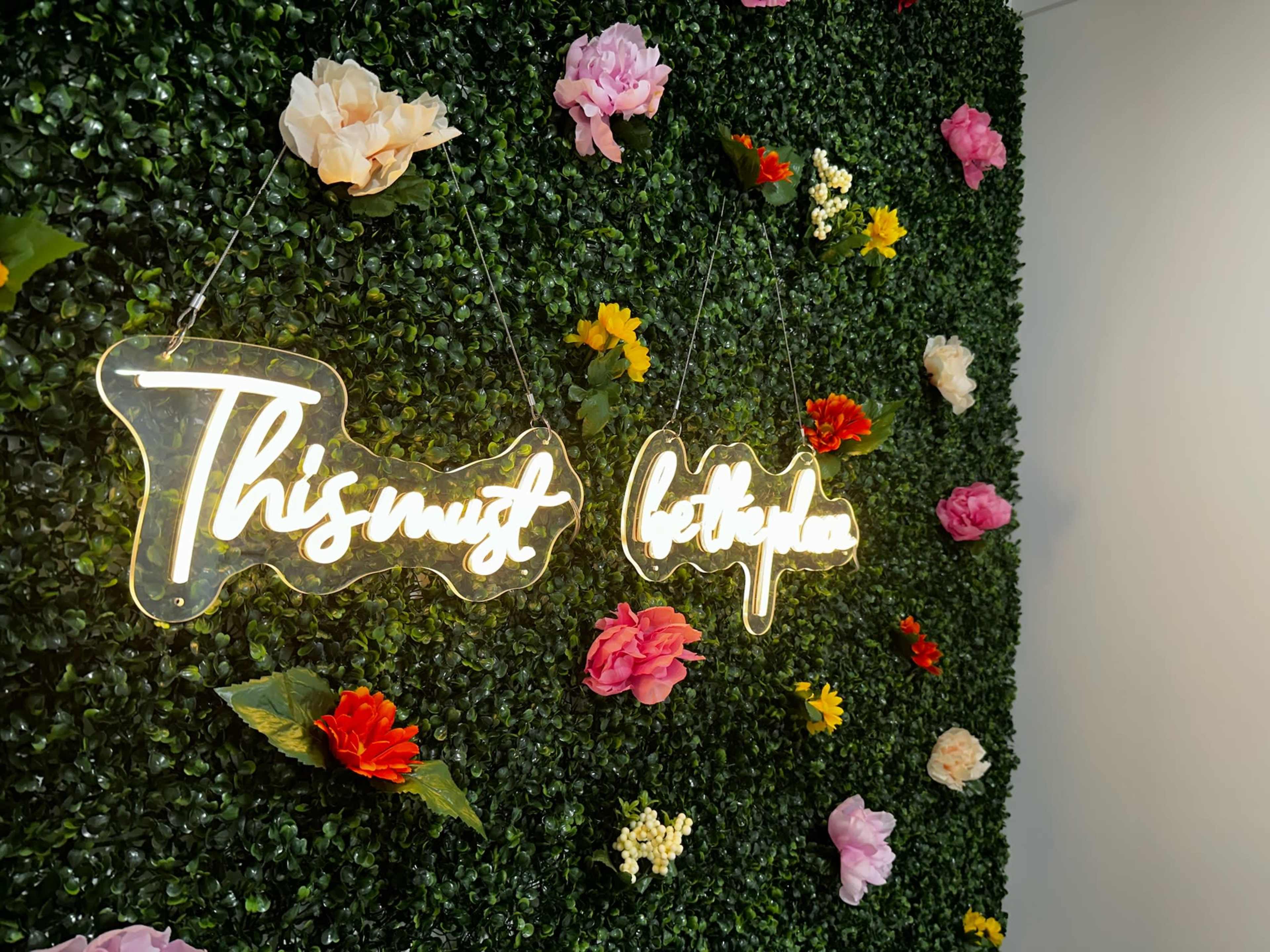 A decorative wall covered in artificial greenery and colorful faux flowers features a glowing sign that reads "This must be the place."