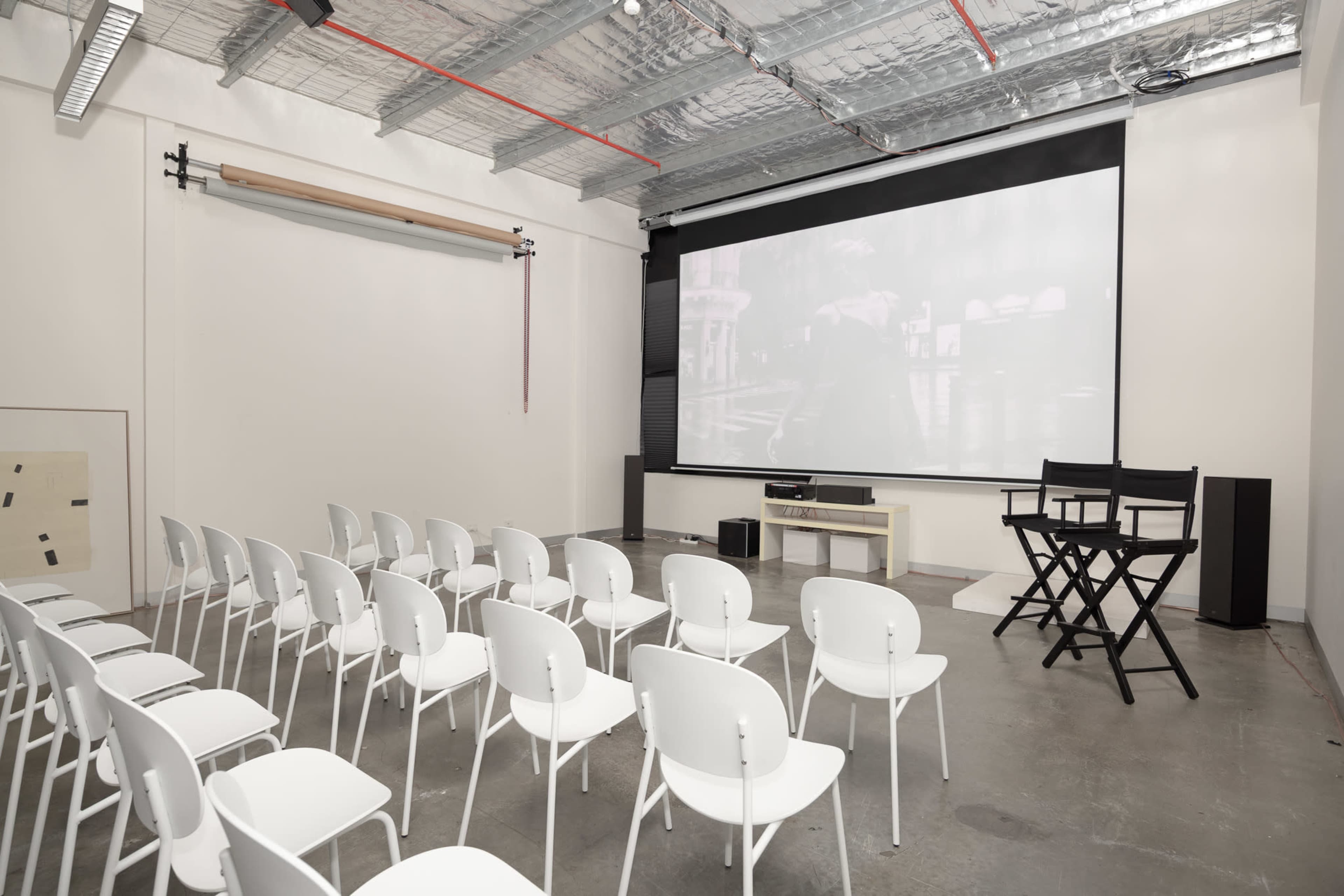 The image shows a minimalist screening room with white chairs arranged in rows facing a large projection screen.